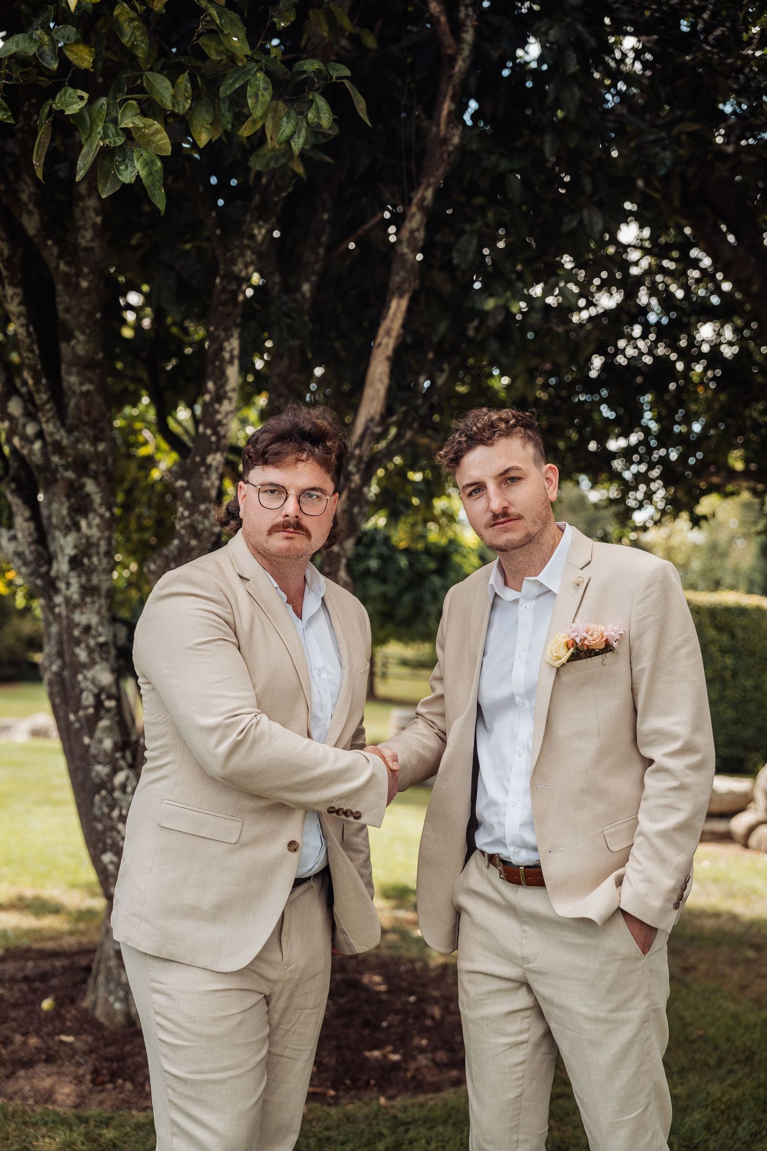 Two grooms in matching beige suits shake hands while posing outdoors near a large tree on their wedding day.