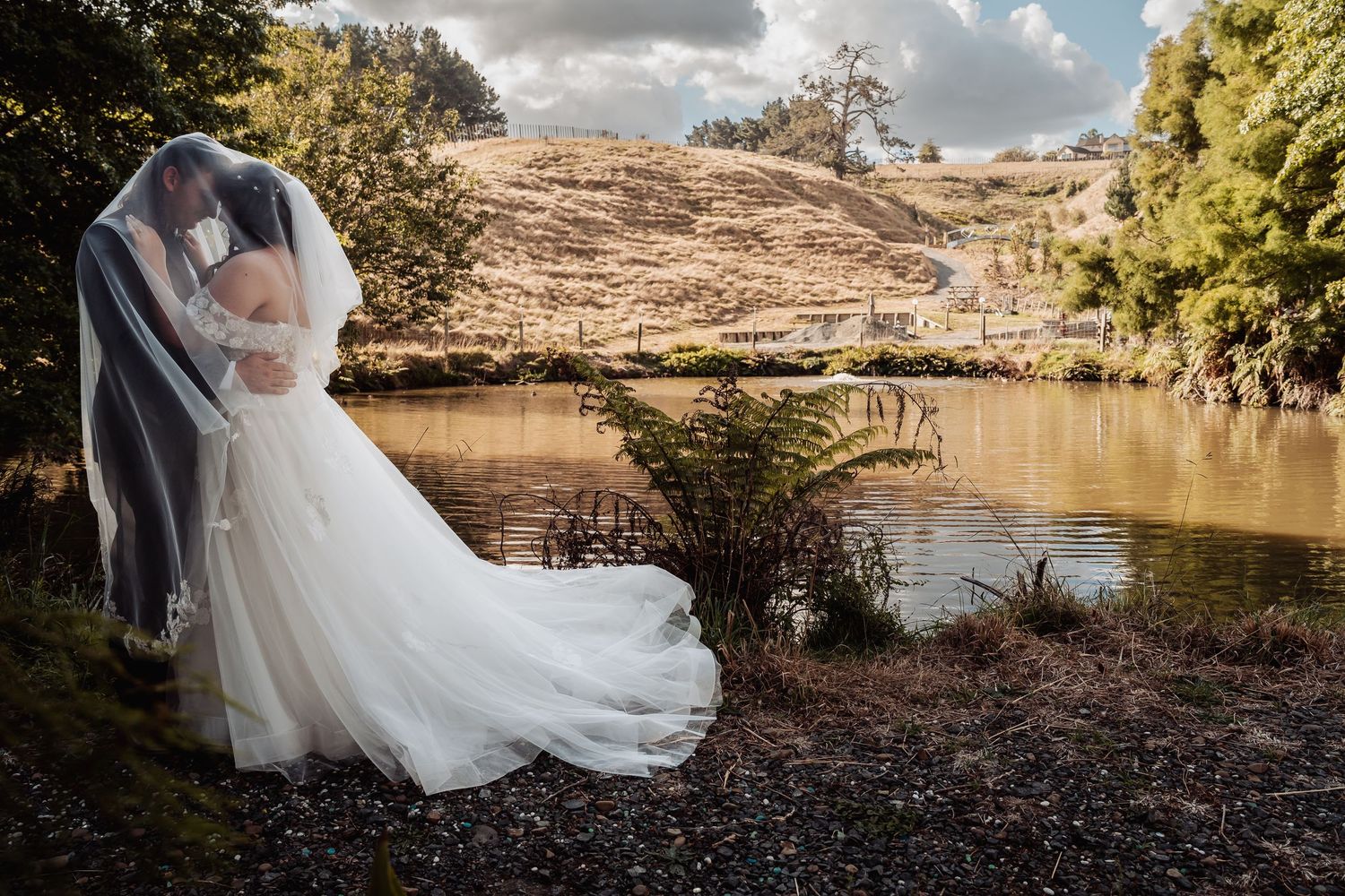 Bride and groom kissing under veil beside tranquil pond with rolling hills in background.