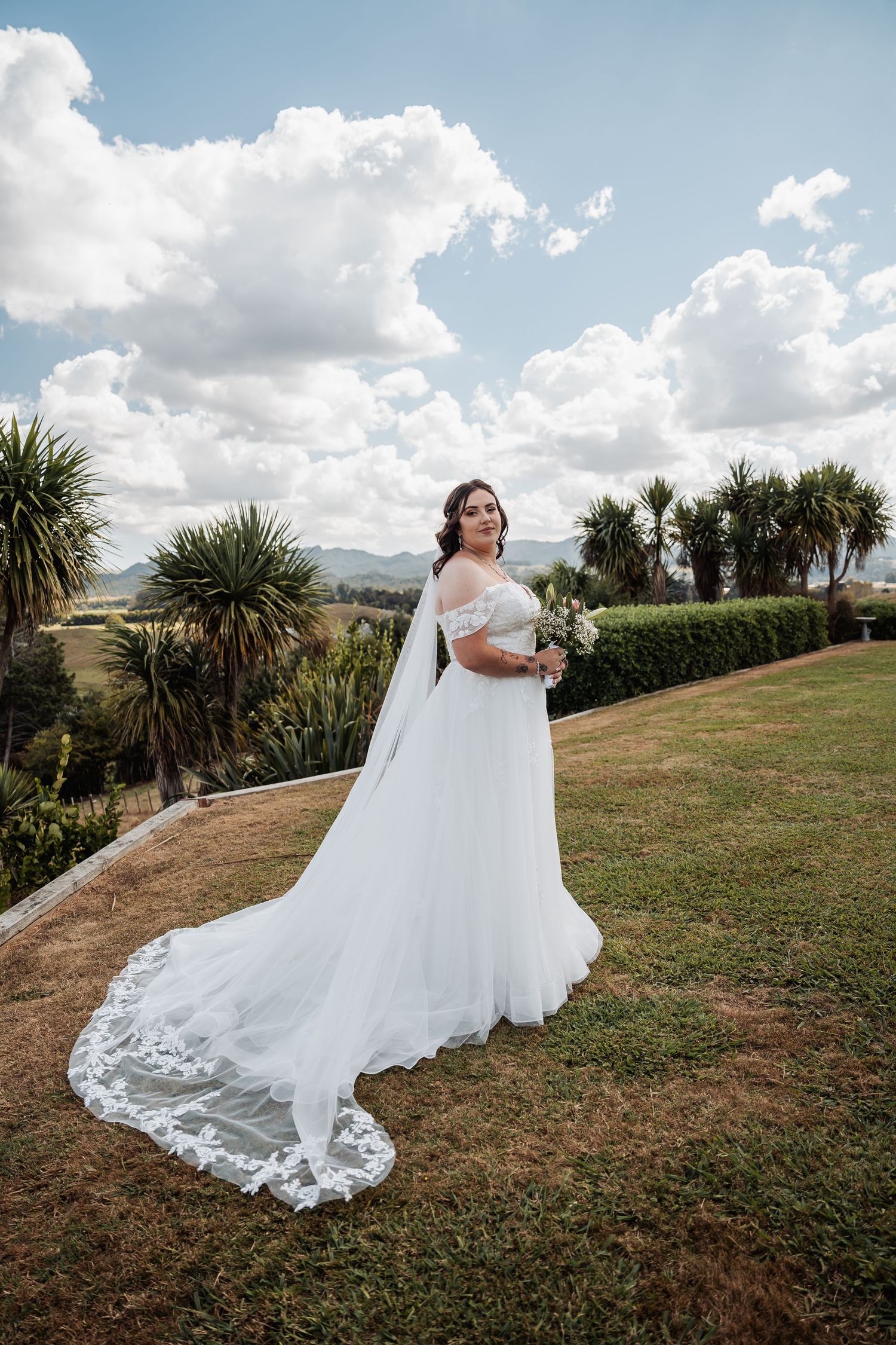 Bride in white off-shoulder gown with lace cape veil holding bouquet in lush garden setting.