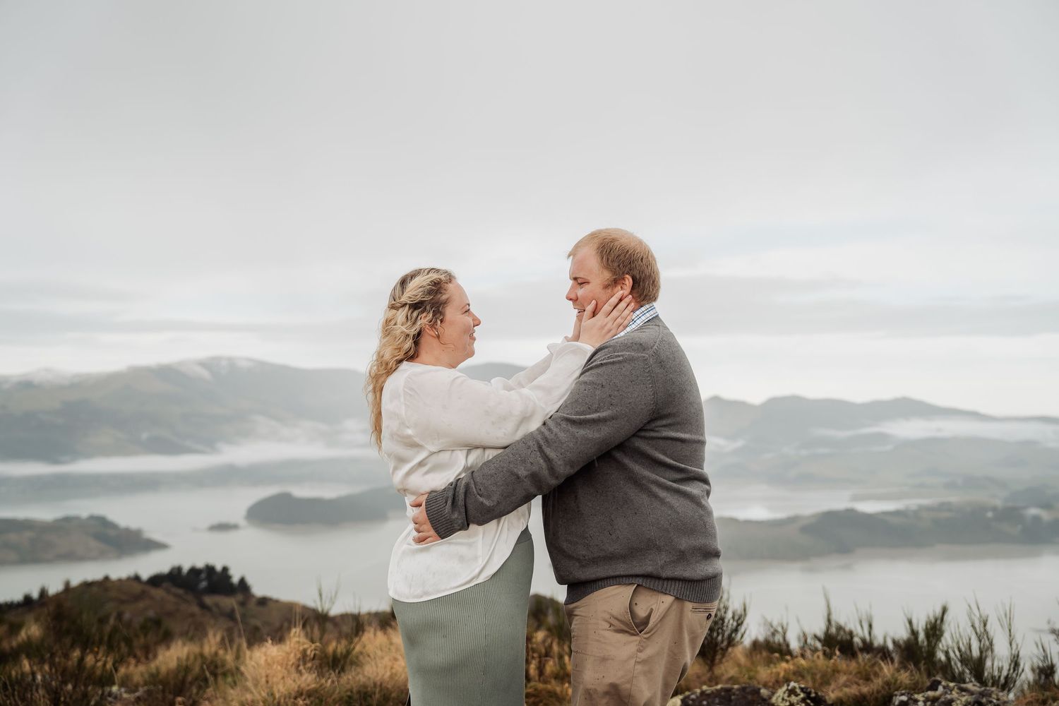 A couple embraces on a misty mountain peak overlooking foggy valleys below during a romantic moment.