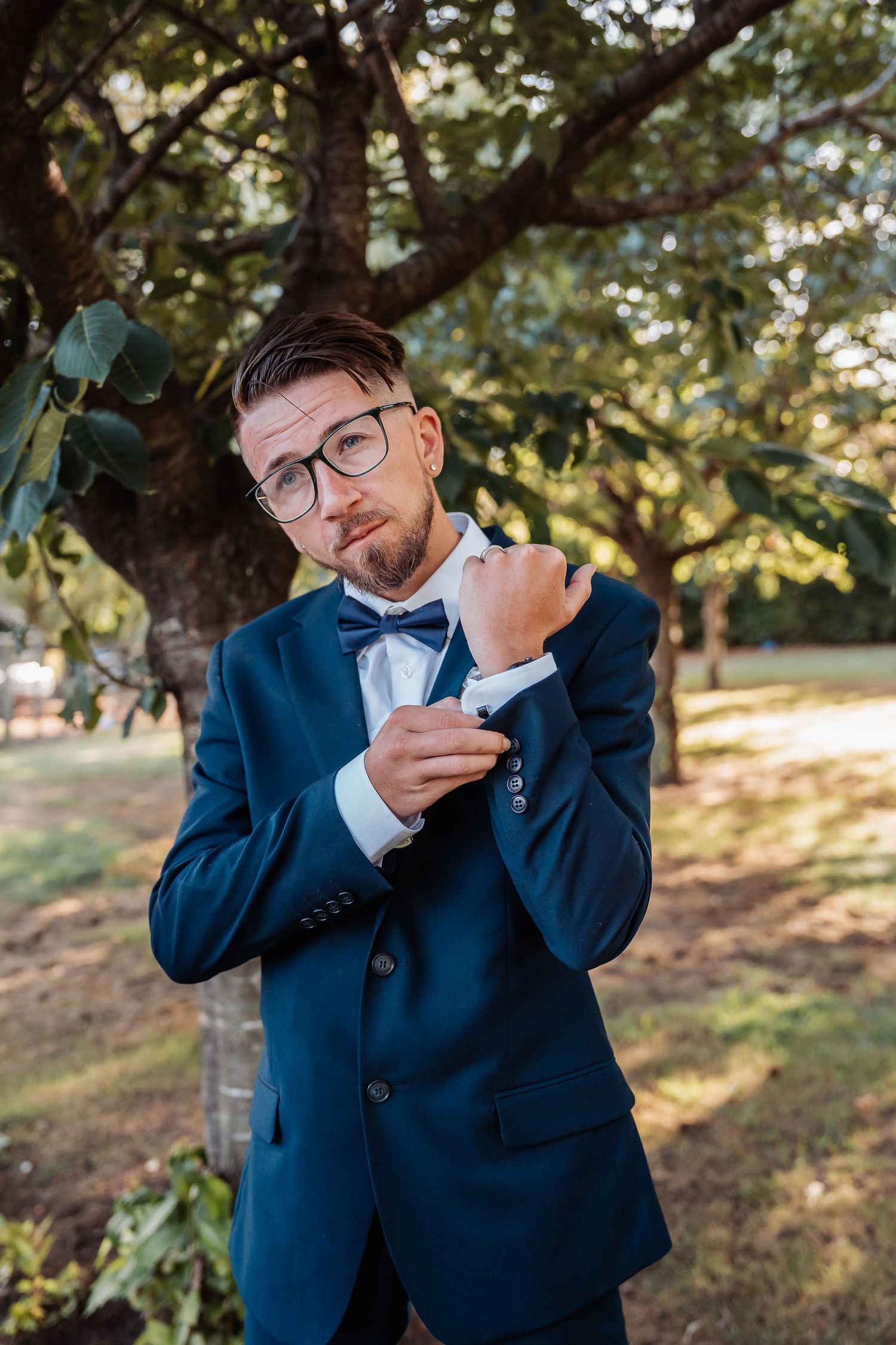 A groom in a navy suit adjusts his bowtie while standing in a garden setting.