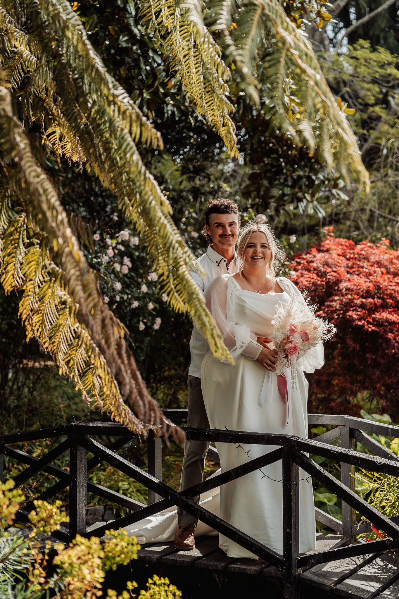 Wedding portrait on garden bridge with hanging ferns and red maple foliage.