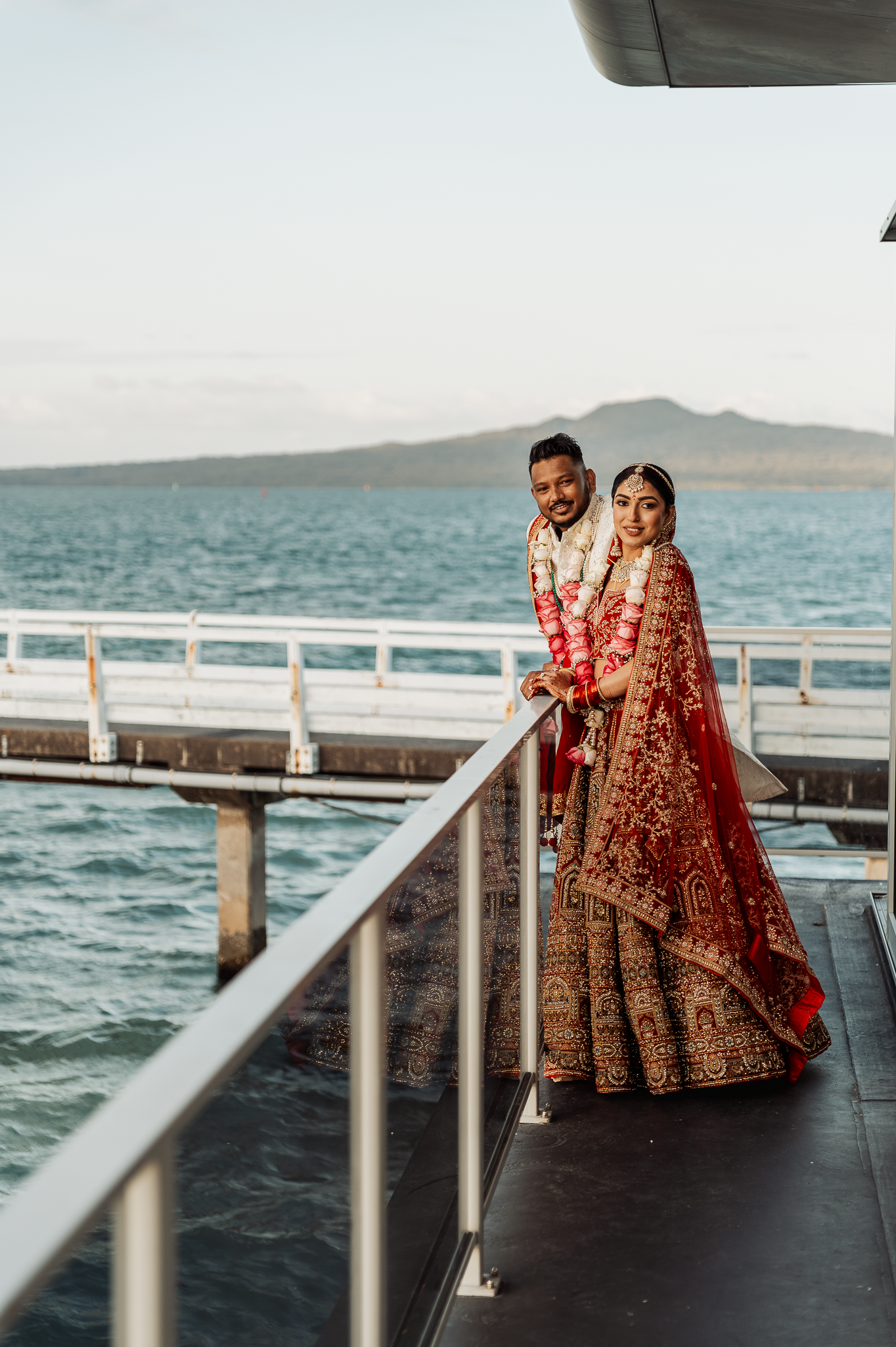 Waterfront wedding photo of couple in traditional Indian attire on pier with ocean view.