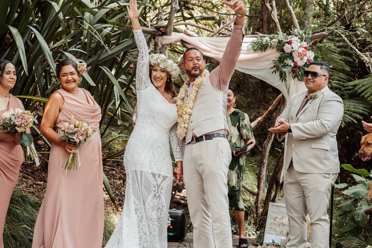 Joyful wedding celebration under a floral arch with bridesmaids in pink dresses.