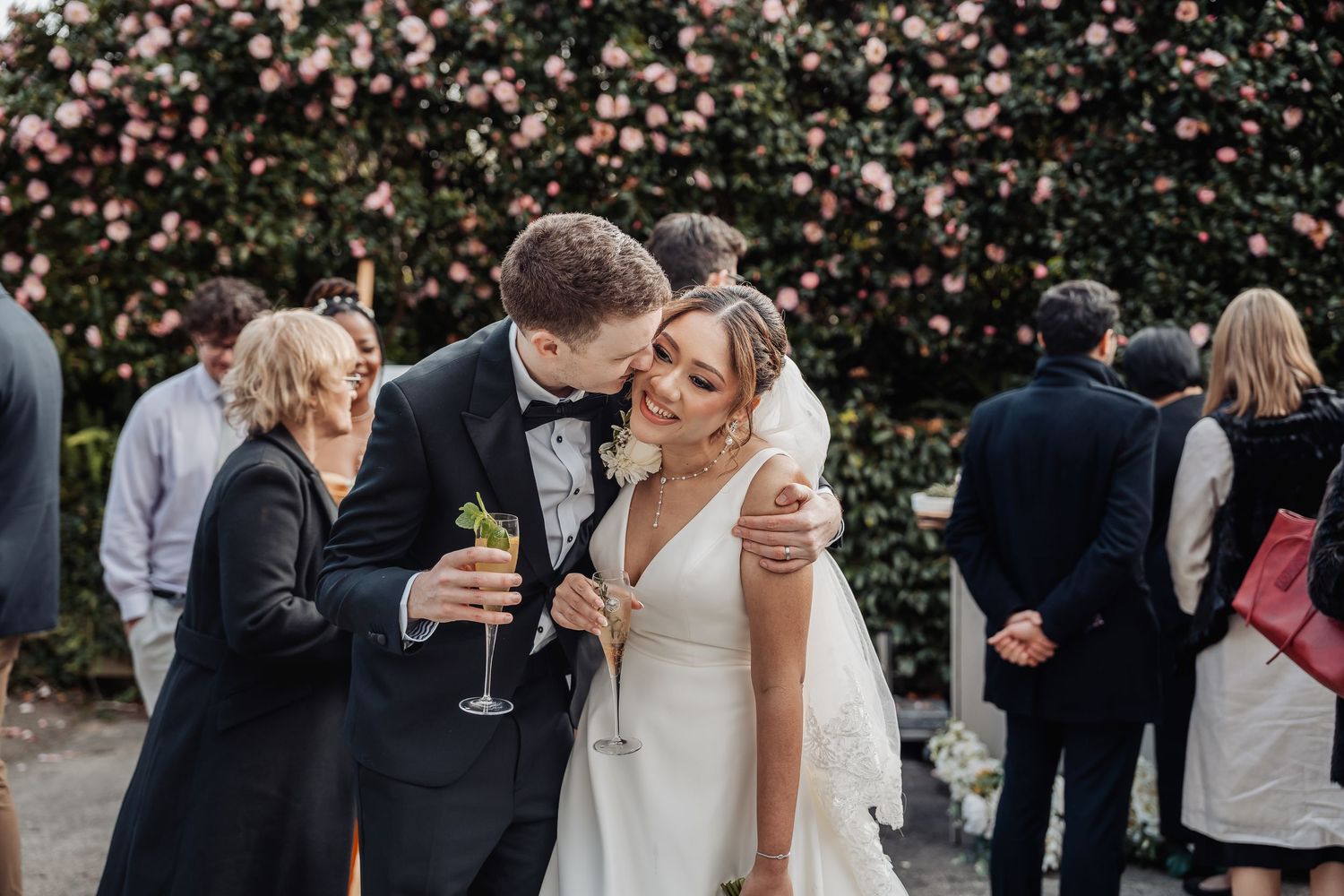 Wedding celebration in progress beneath flowering pink and white cherry blossom trees in a garden setting.