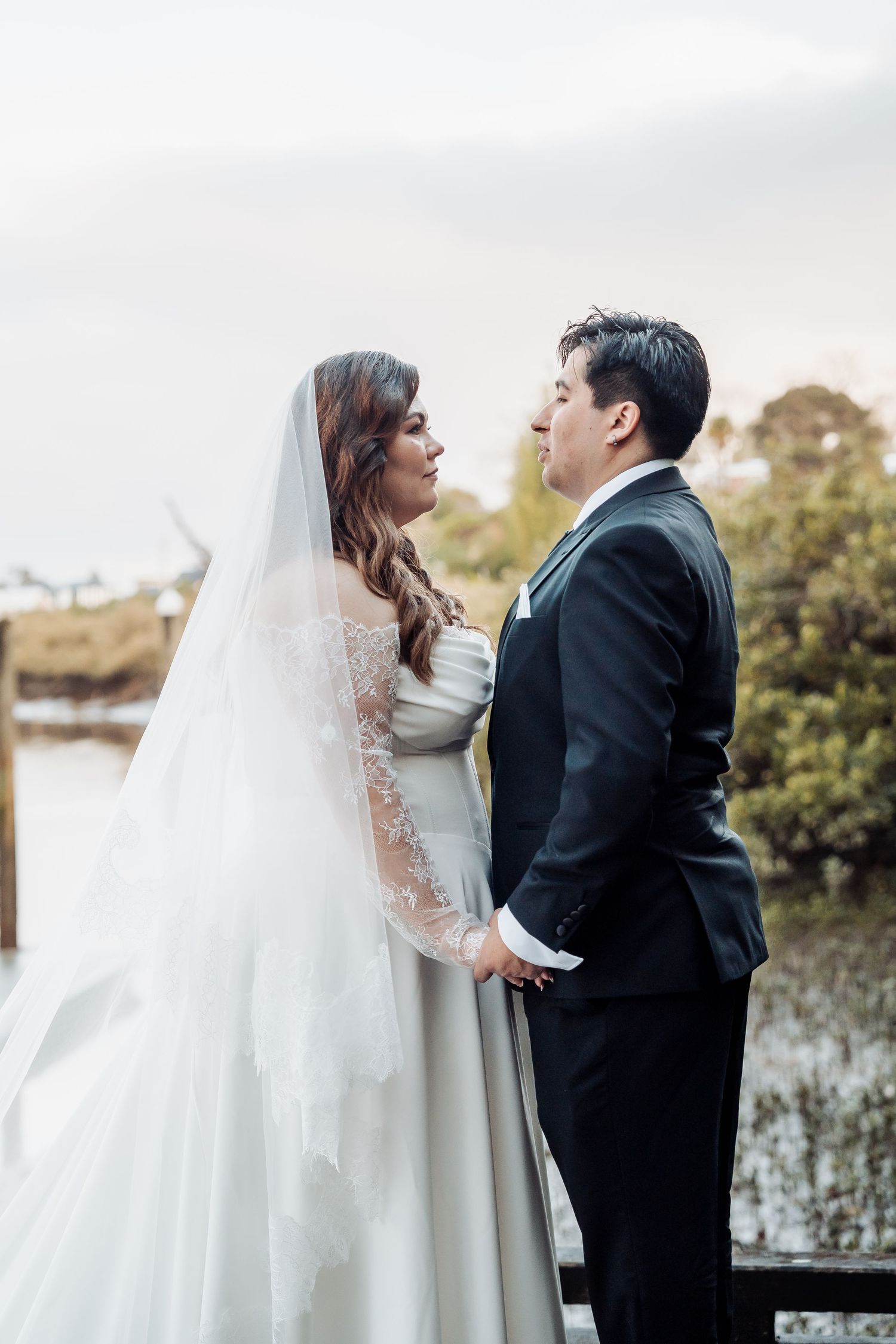 A couple shares an intimate moment during their outdoor wedding ceremony by a lake, with the bride in a long-sleeved lace gown.