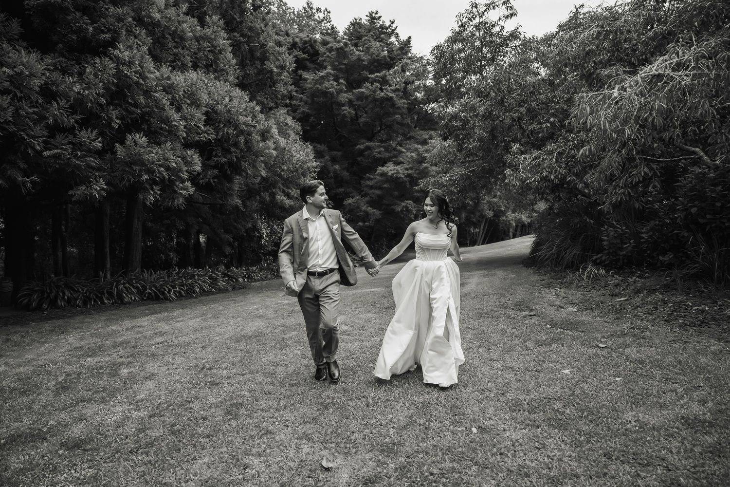 Black and white photo of smiling newlyweds holding hands walking across lush green lawn.