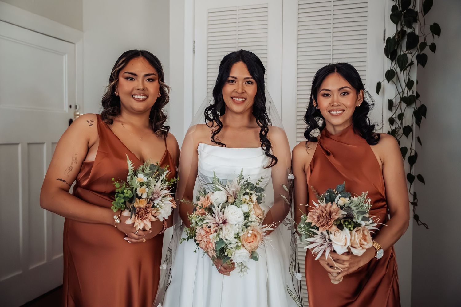 Bride in white gown with two bridesmaids in rust satin dresses holding bouquets of pink and white flowers.