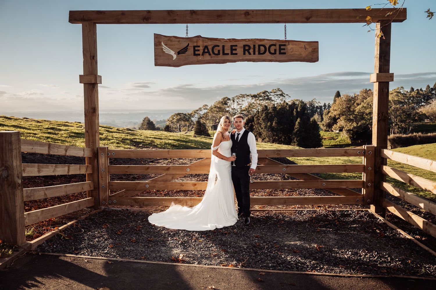 Bride and groom pose under rustic Eagle Ridge wooden sign with scenic mountain views at outdoor wedding venue.