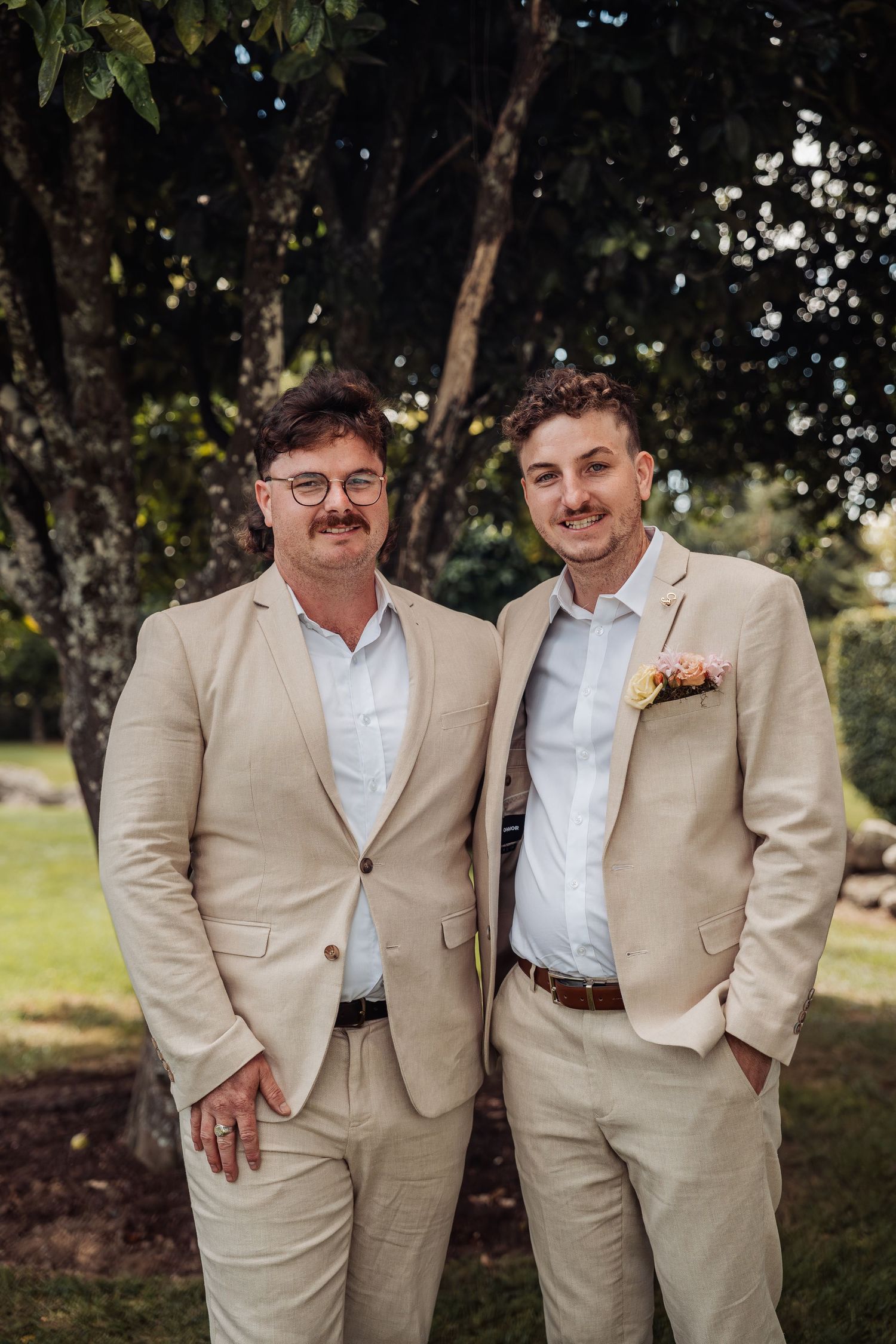 Two men in matching beige suits smiling for a formal portrait outdoors at a wedding.