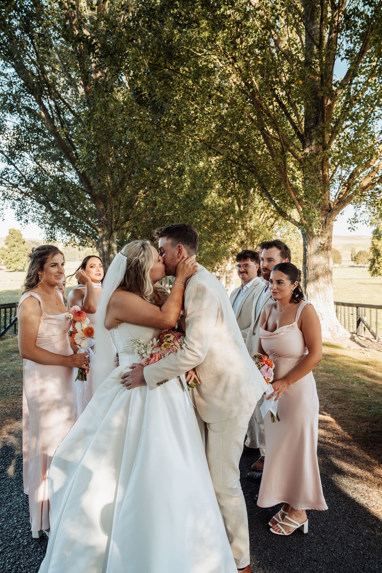 Bride and groom kiss surrounded by bridesmaids and groomsmen under tall trees at wedding.