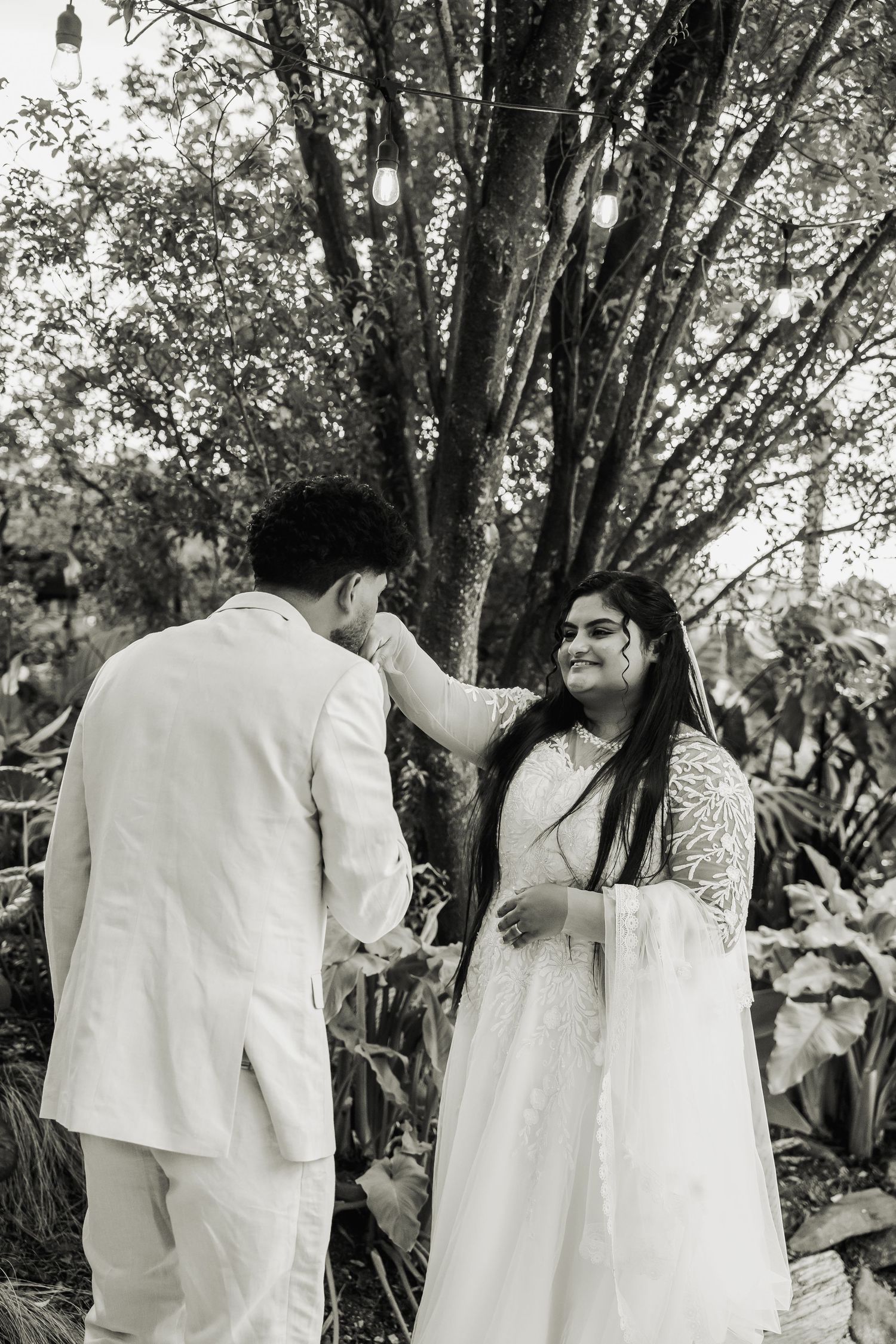 Black and white photo of newlyweds sharing tender moment under tree with string lights.