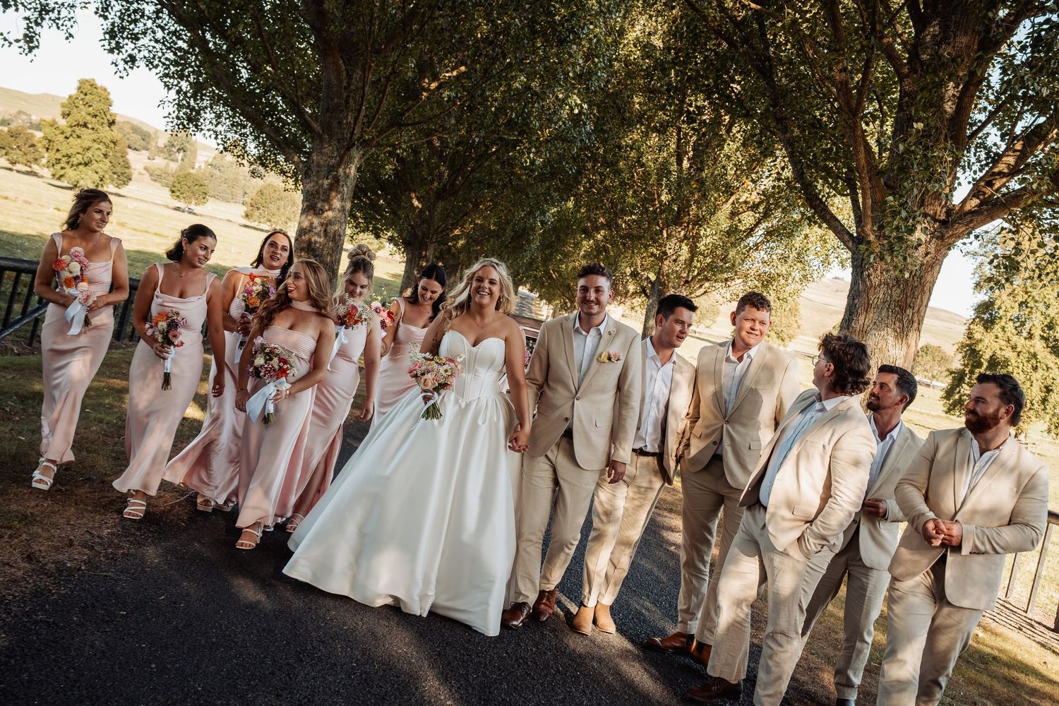 A wedding party in blush bridesmaid dresses and tan suits walks together under large trees outdoors.