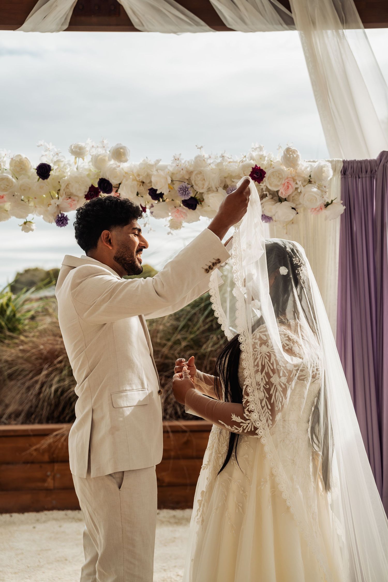 Groom lifts bride's veil during romantic outdoor wedding ceremony under floral decorated arch.