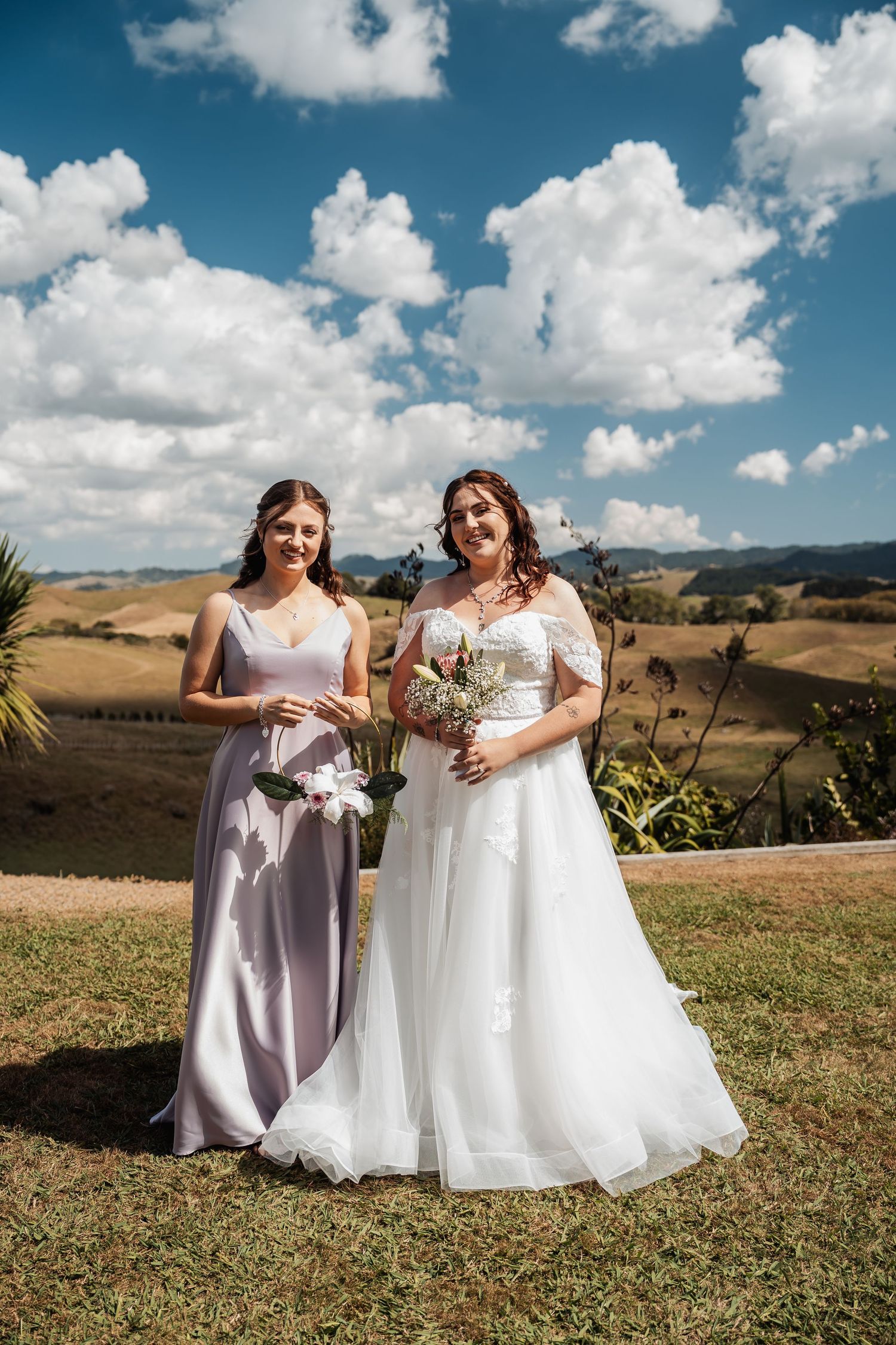 Bride in white off-shoulder gown poses with bridesmaid in lavender dress holding bouquets outdoors.
