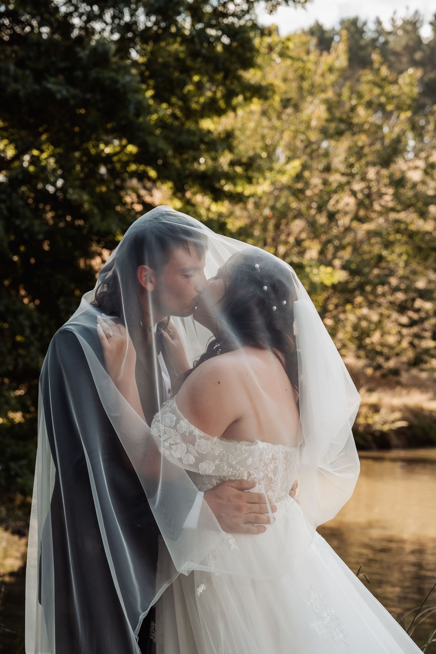 Bride and groom sharing a kiss under a flowing veil near a sunlit forest pond.