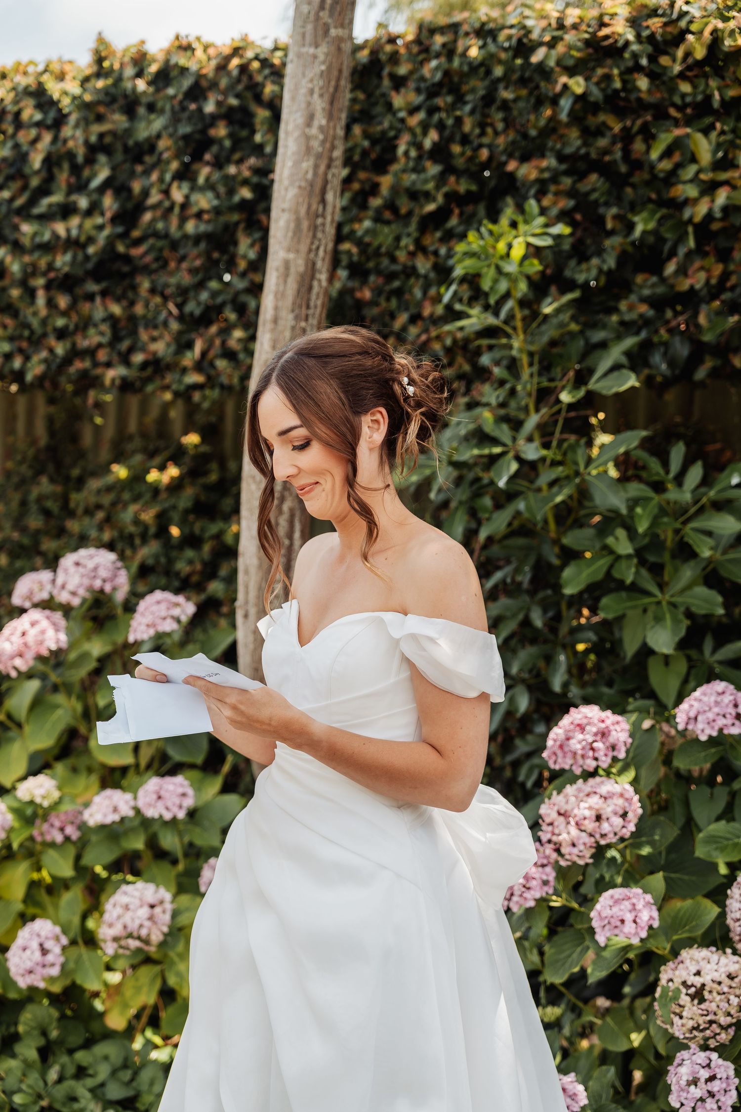 Smiling bride in off-shoulder white gown reading vows beside pink hydrangeas in a lush garden.