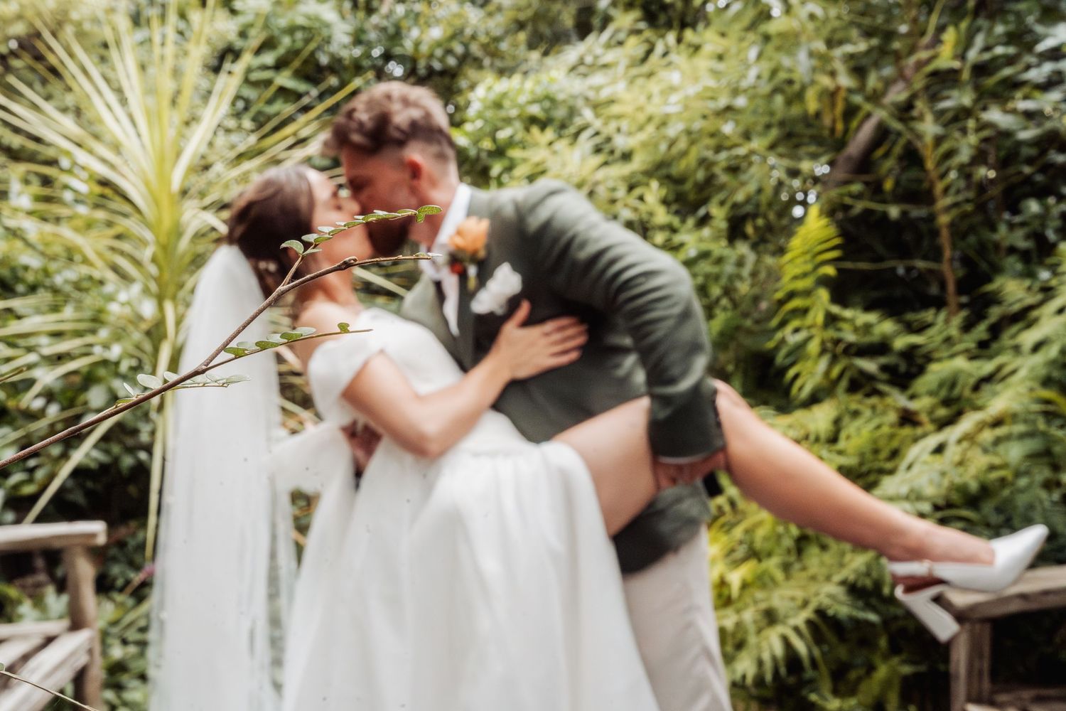 Groom dipping and kissing bride in white gown and veil among lush tropical garden greenery.