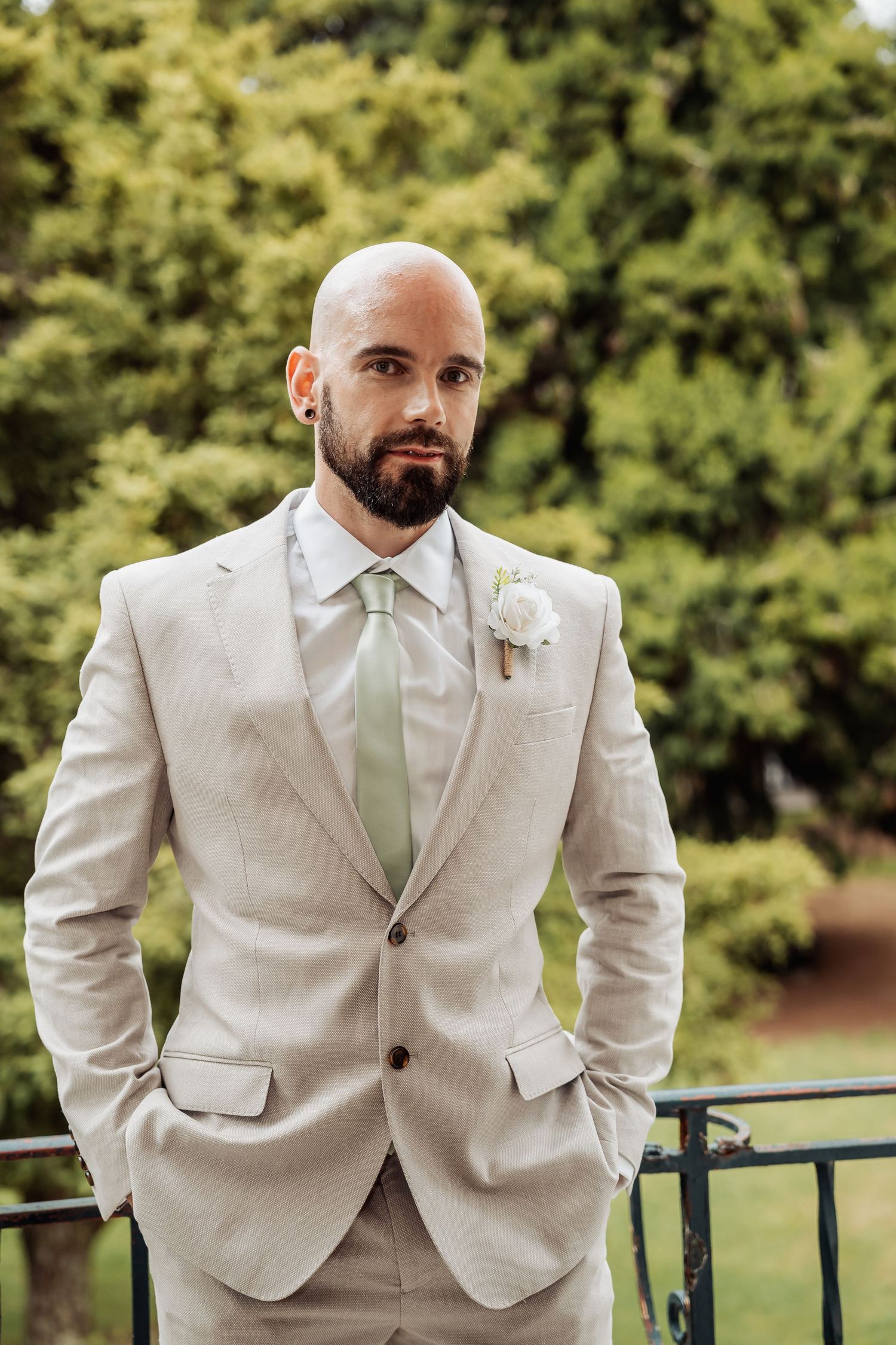 Bald groom with beard wearing beige suit and sage green tie with white boutonniere outdoors.
