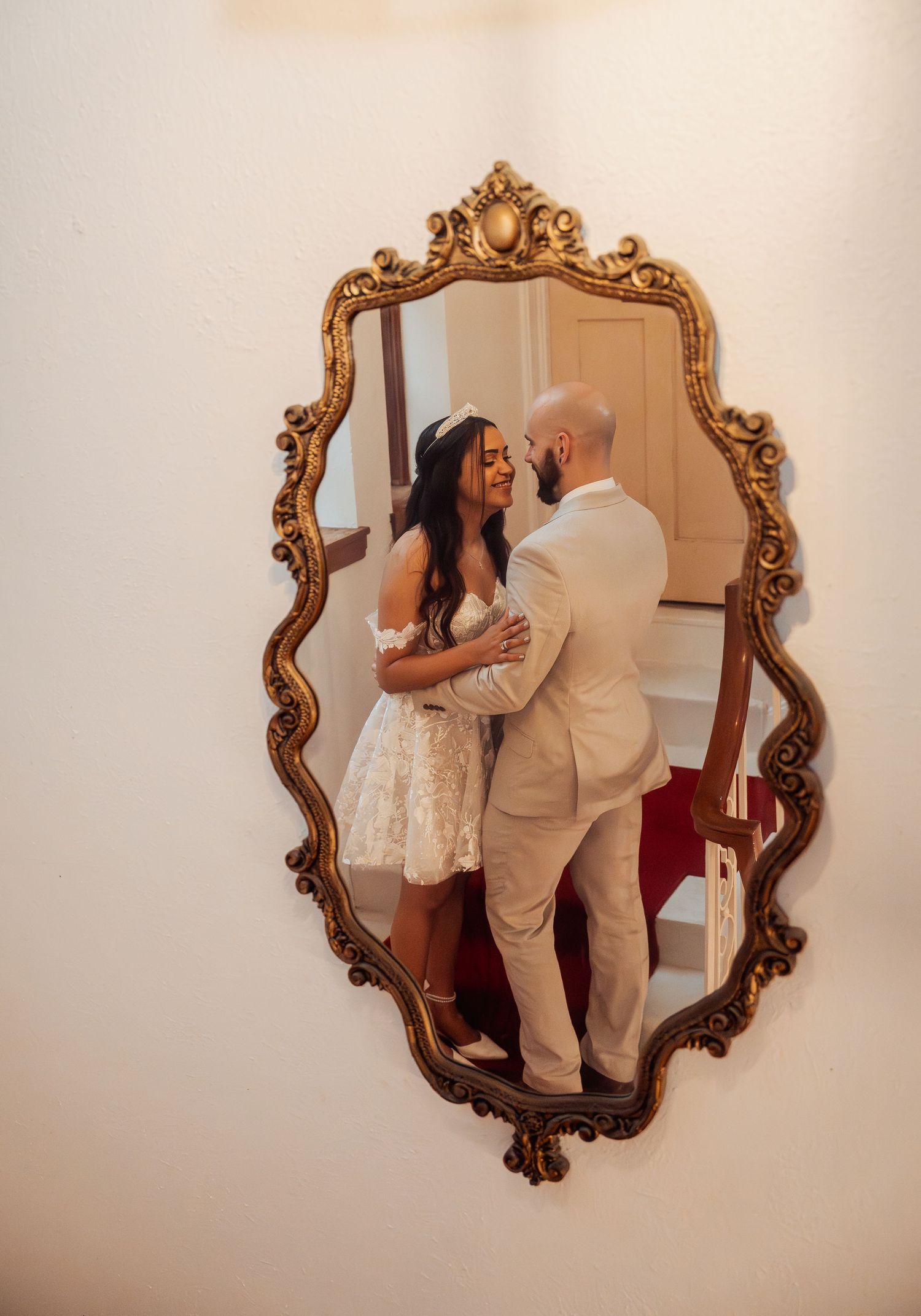 Bride and groom share a tender embrace reflected in an ornate gold vintage mirror on a white wall.