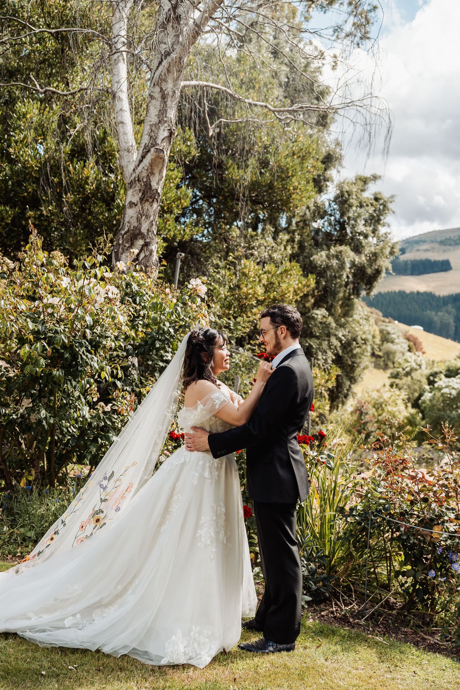 Bride and groom embracing in a scenic garden with rolling hills, bride wearing a floral embroidered veil.