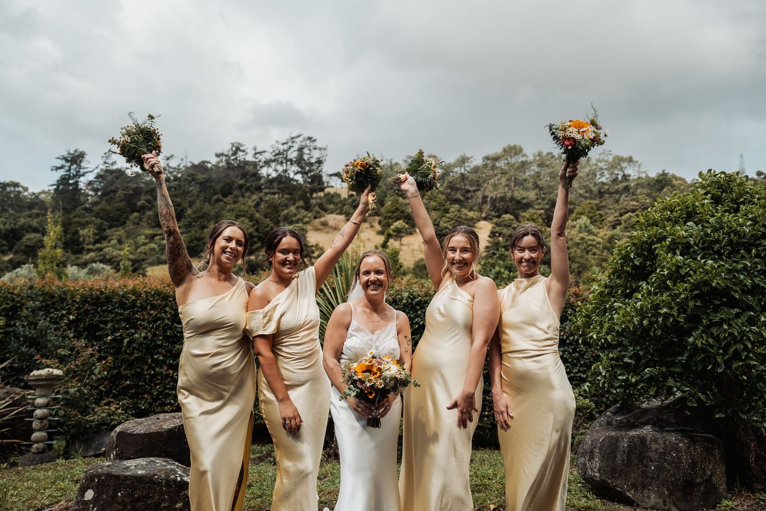 Bride with four bridesmaids in gold dresses raising bouquets outdoors after wedding ceremony.