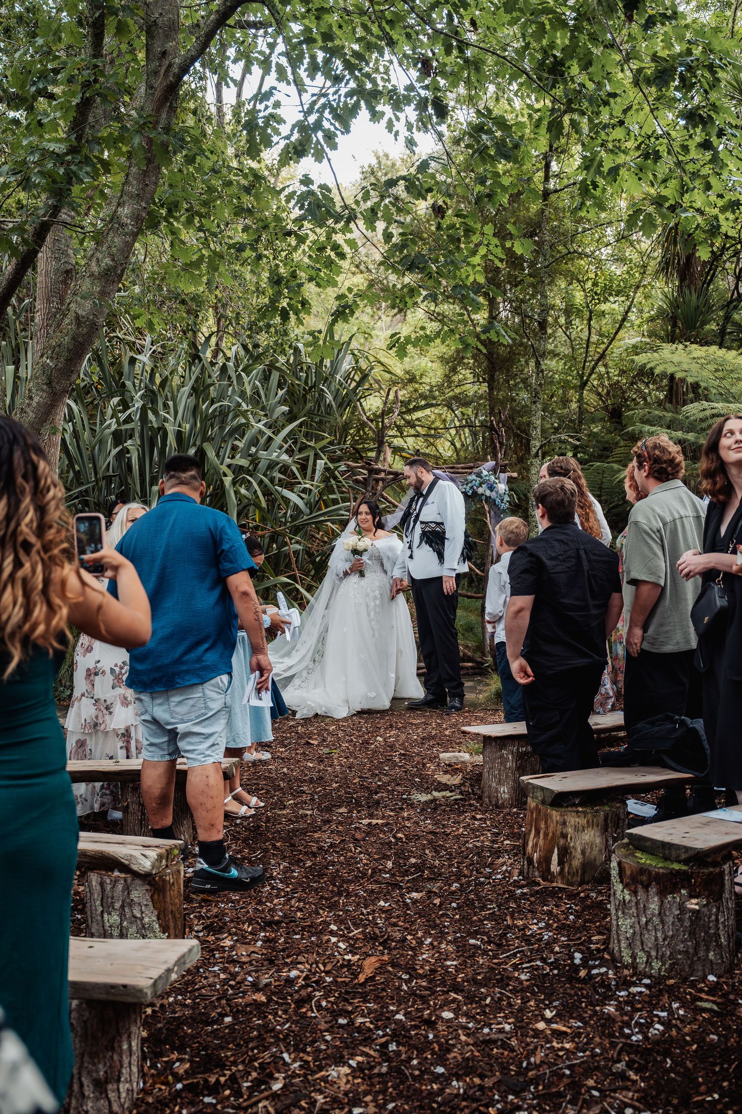 Newlyweds walk down a rustic woodland aisle lined with tree stump seats after their wedding ceremony.