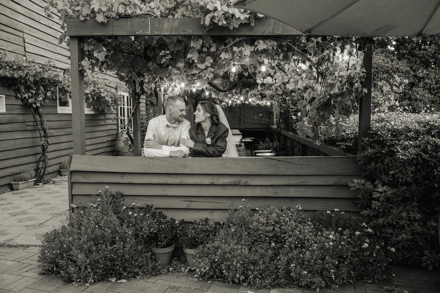 Black and white photo of a couple sharing an intimate moment on a bench beneath a vine-covered pergola.