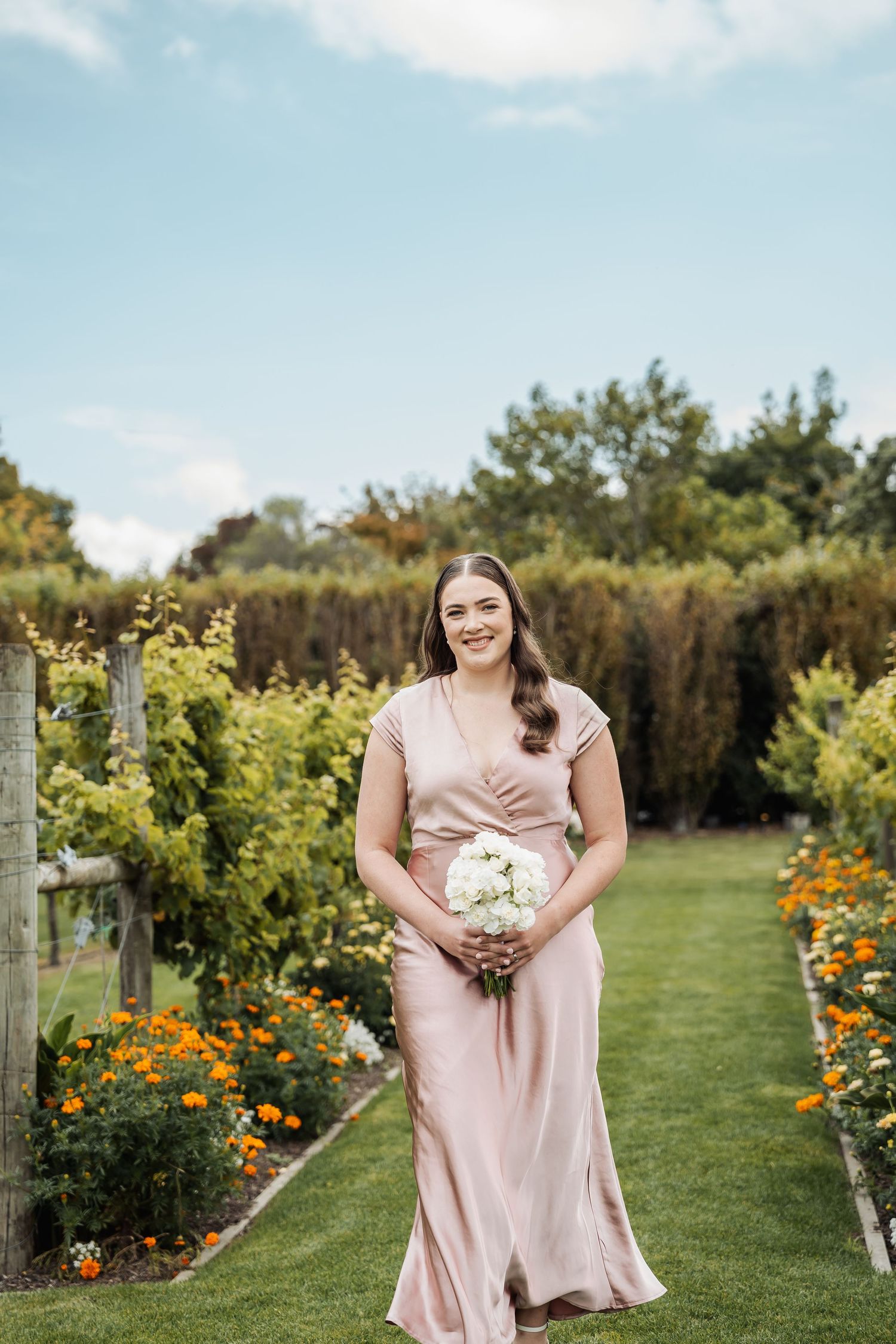 Smiling bridesmaid in blush satin dress holds white bouquet while standing in a colorful garden path.