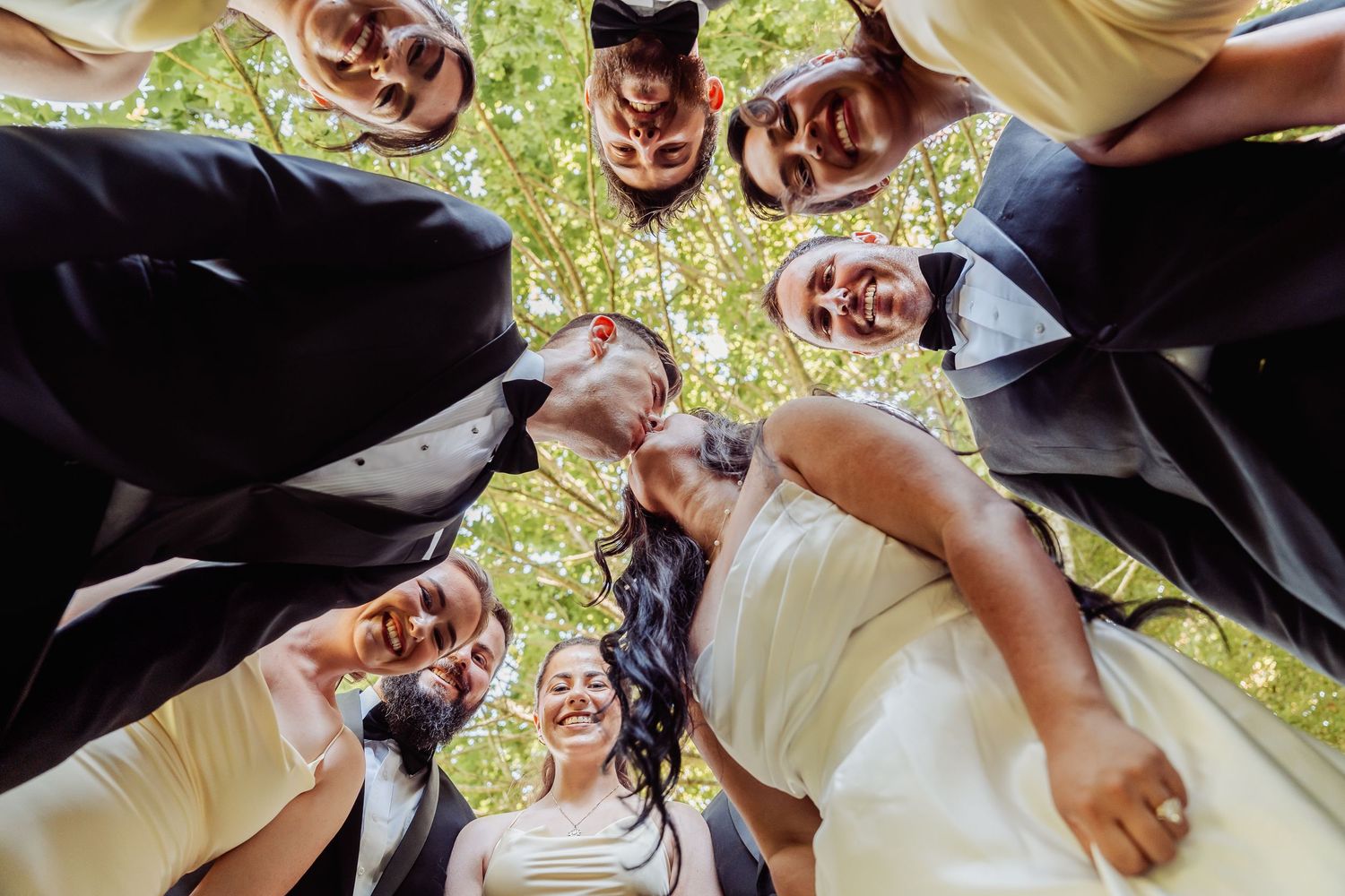 Wedding party forms a circle huddle looking down at smiling bride in white dress outdoors.