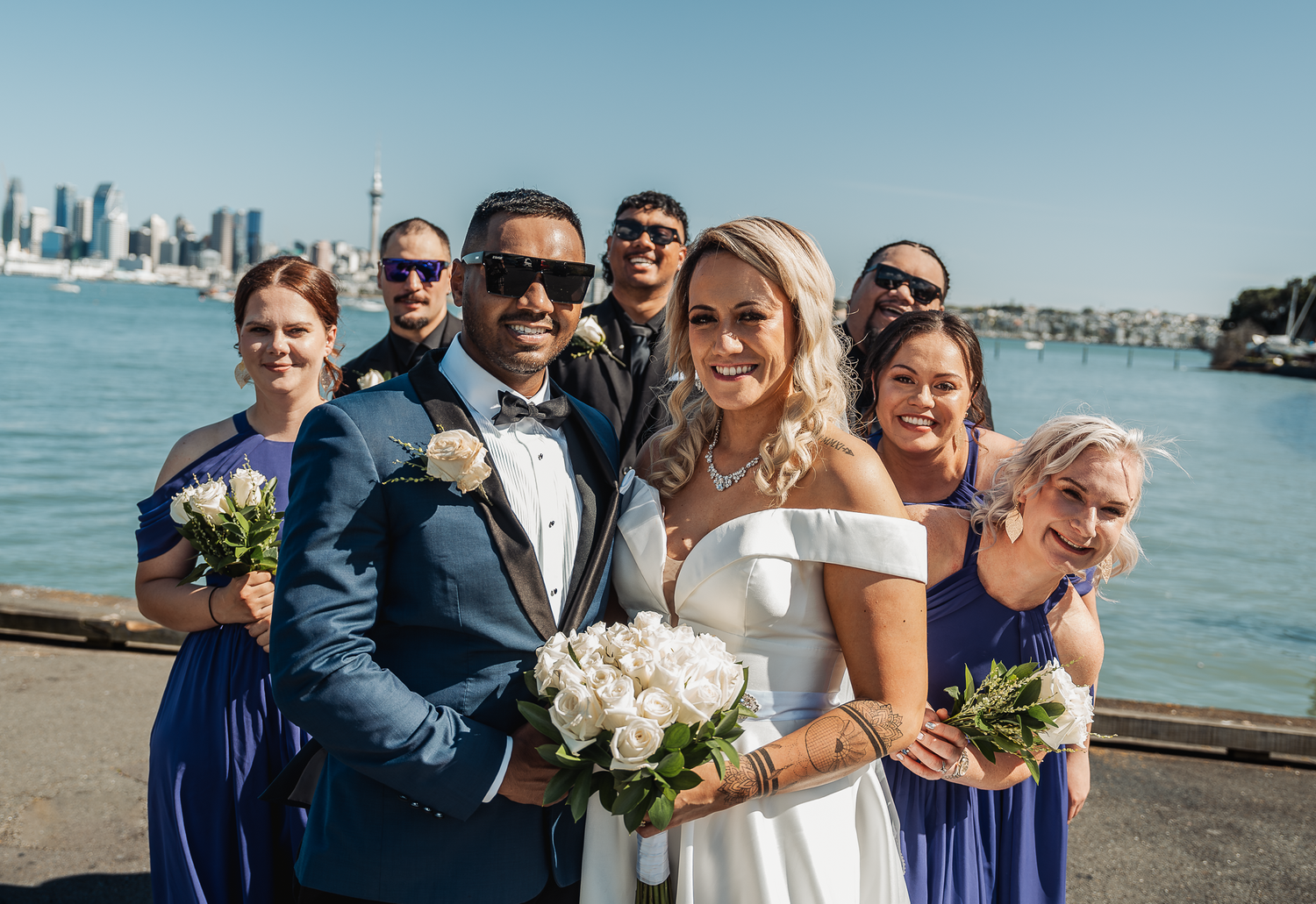 Wedding party poses on waterfront dock with city skyline backdrop and bridesmaids in navy blue dresses.