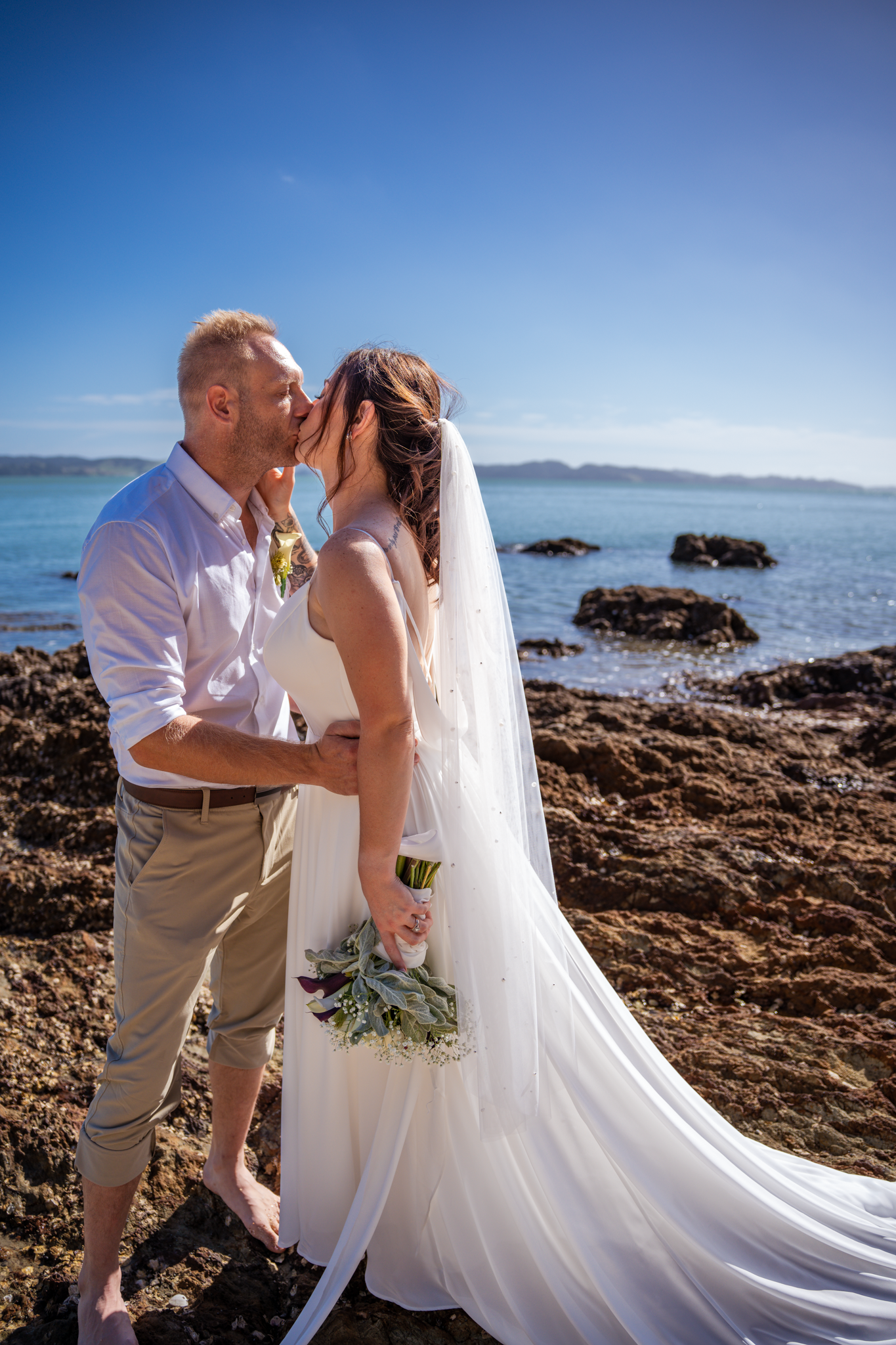 Romantic beach wedding portrait with white flowing dress against coastal rocks and blue water.