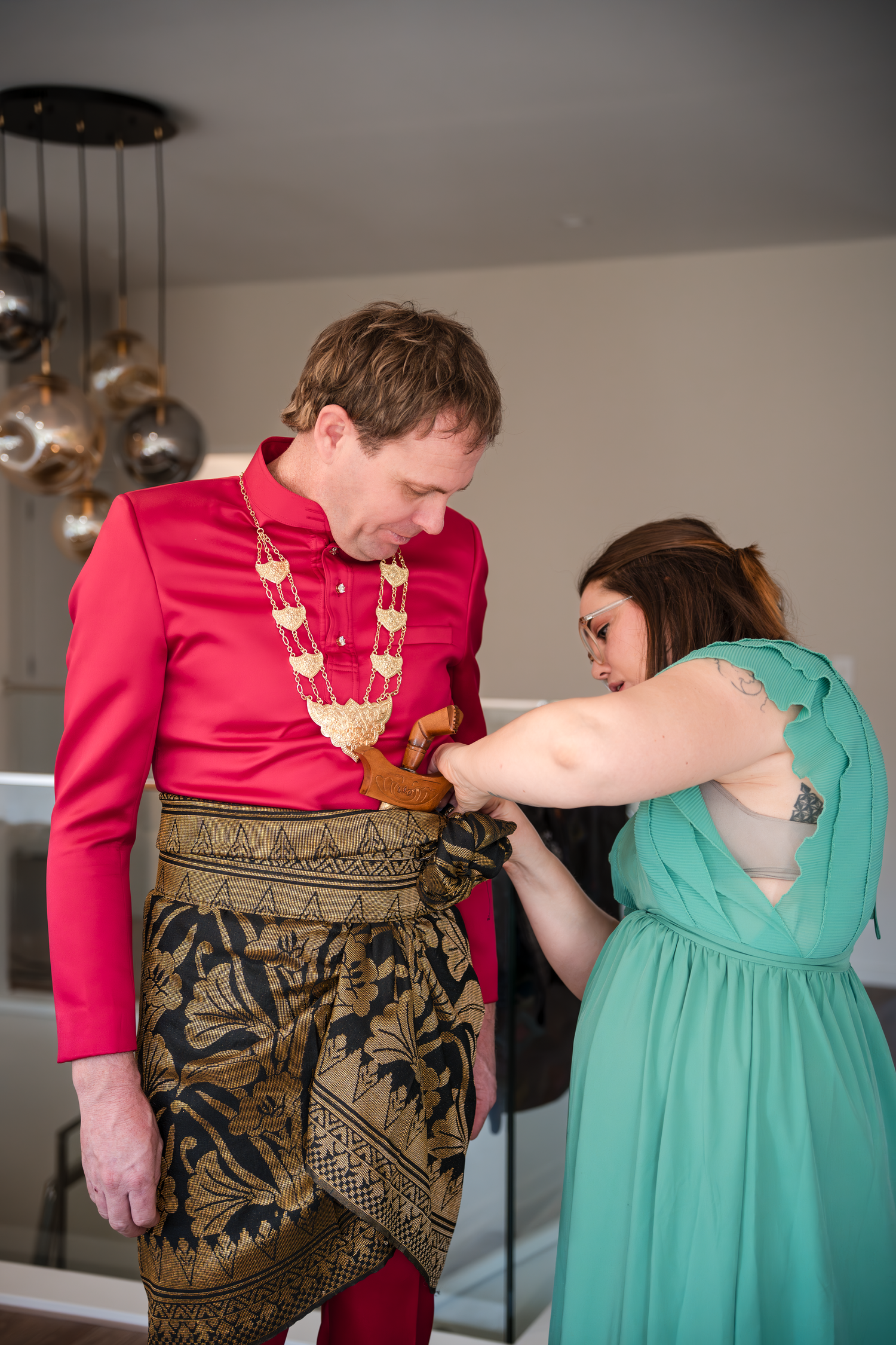 A couple getting ready for a wedding, with one in a red ceremonial jacket and gold patterned sarong.