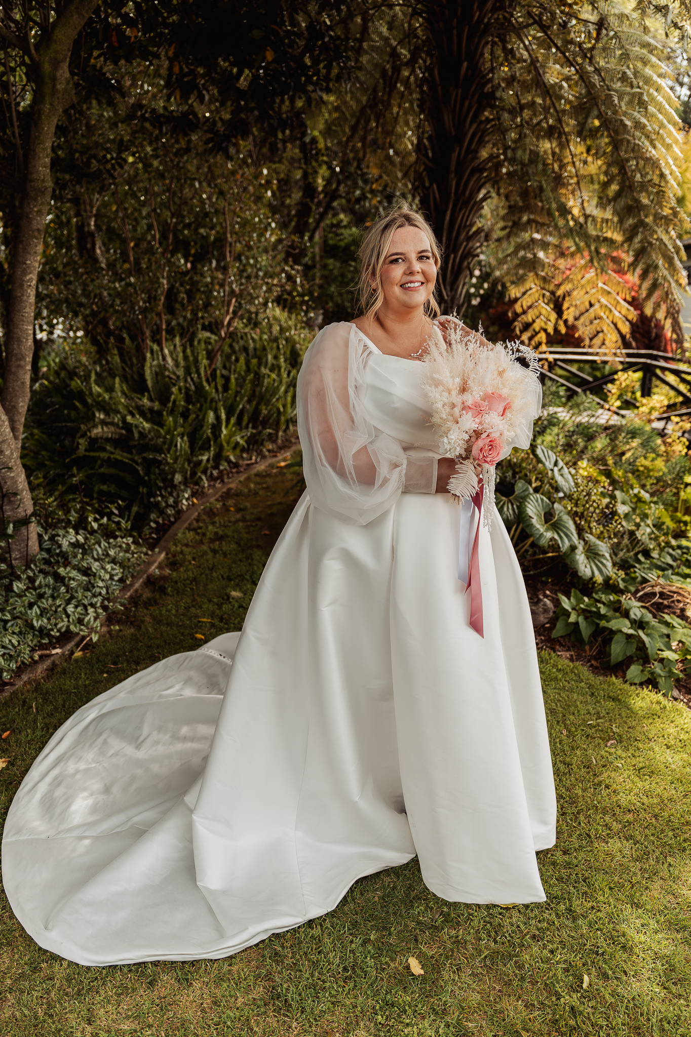 Bride in elegant white gown with flowing train holds pink bouquet in garden setting.