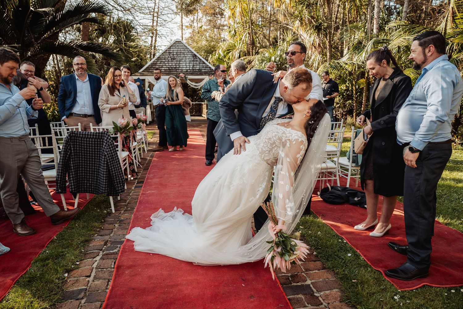 Wedding ceremony guests seated on white chairs along red carpet aisle in garden setting.