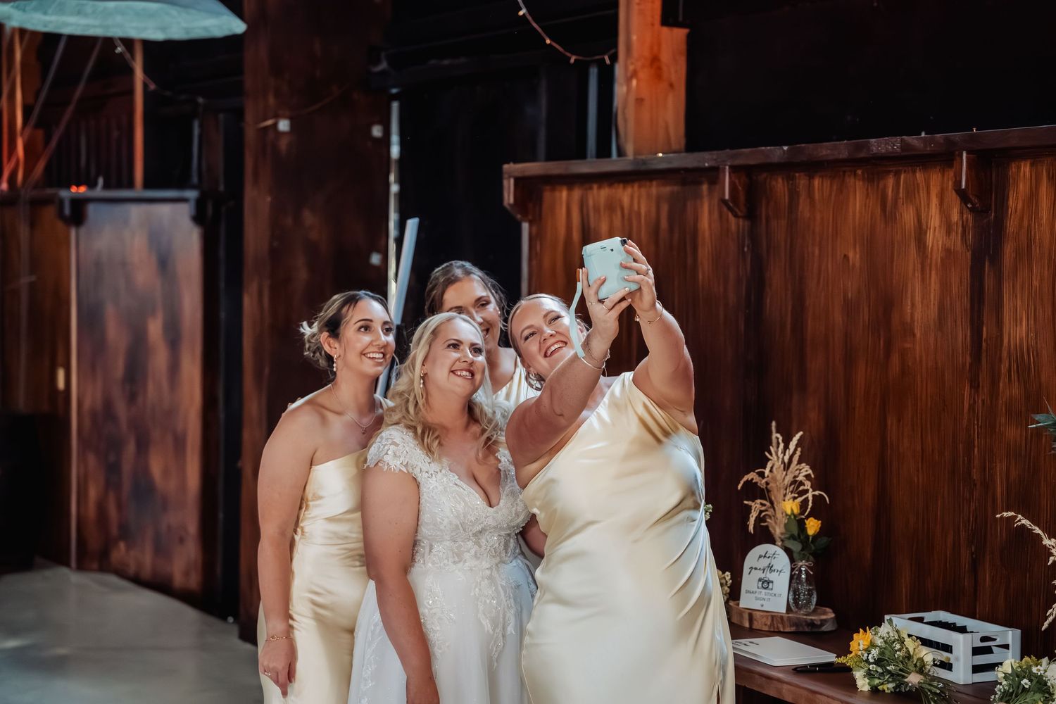 Bride in lace gown and bridesmaids in ivory dresses taking a selfie inside a rustic barn wedding venue.
