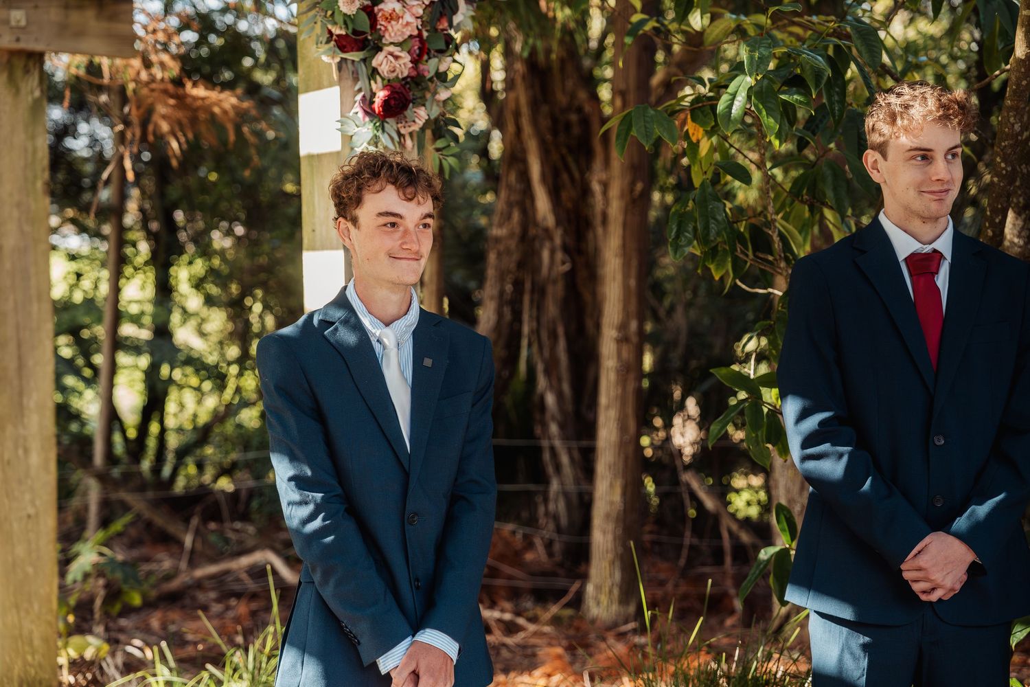 Two young groomsmen in navy suits standing at outdoor wedding ceremony altar.