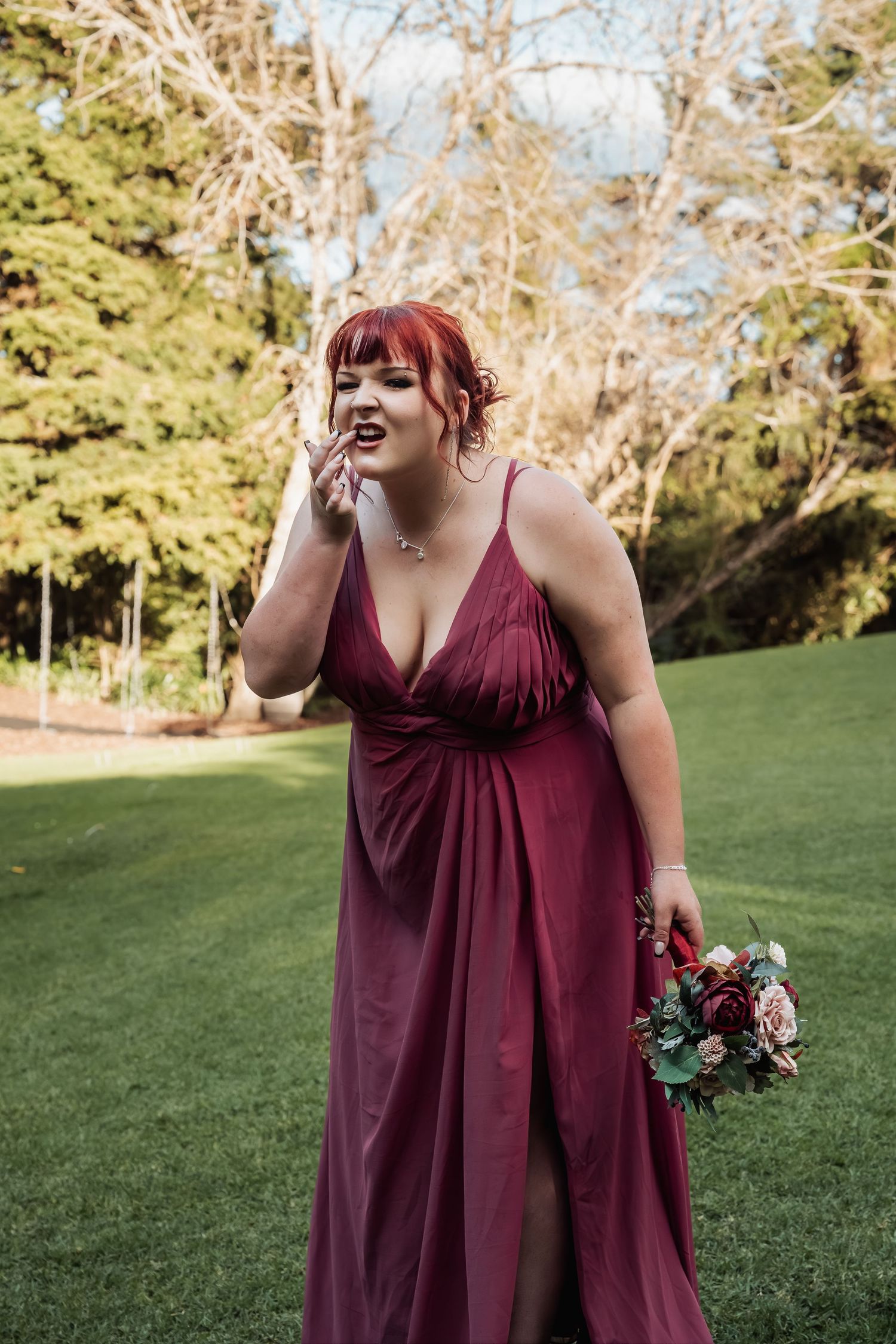 Bridesmaid in burgundy gown holding bouquet outdoors at wedding venue.