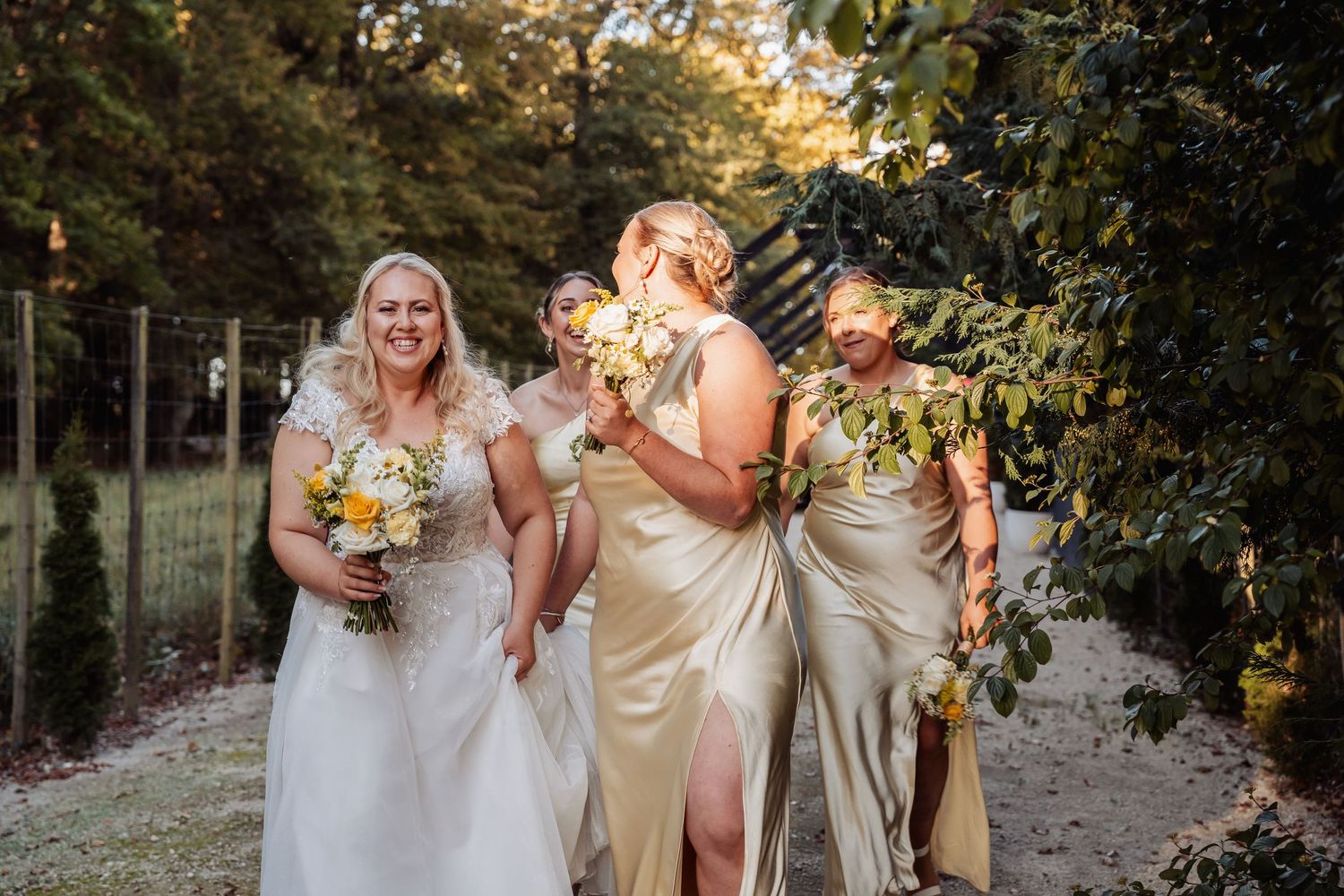 Bride in white lace gown embracing bridesmaids in gold satin dresses in garden setting.
