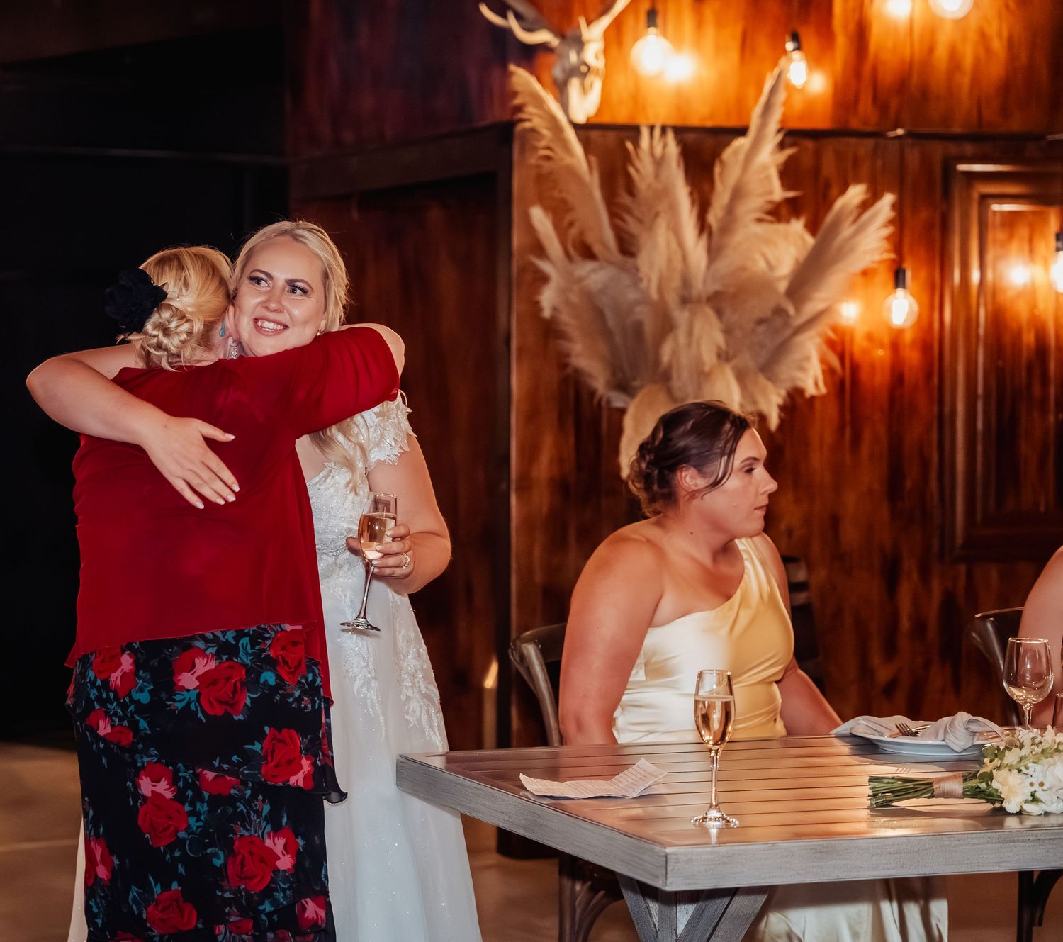 Wedding guest in red floral dress hugging bride during indoor reception with warm string lights.