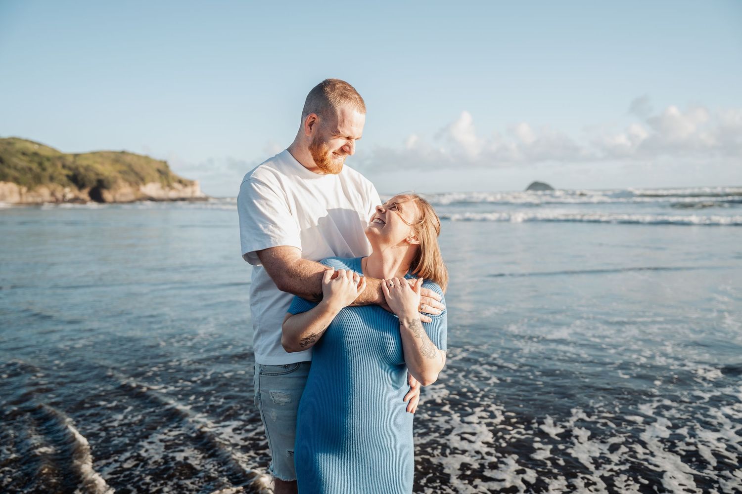 Two people embrace while standing in shallow ocean water against a scenic coastal landscape with cliffs.