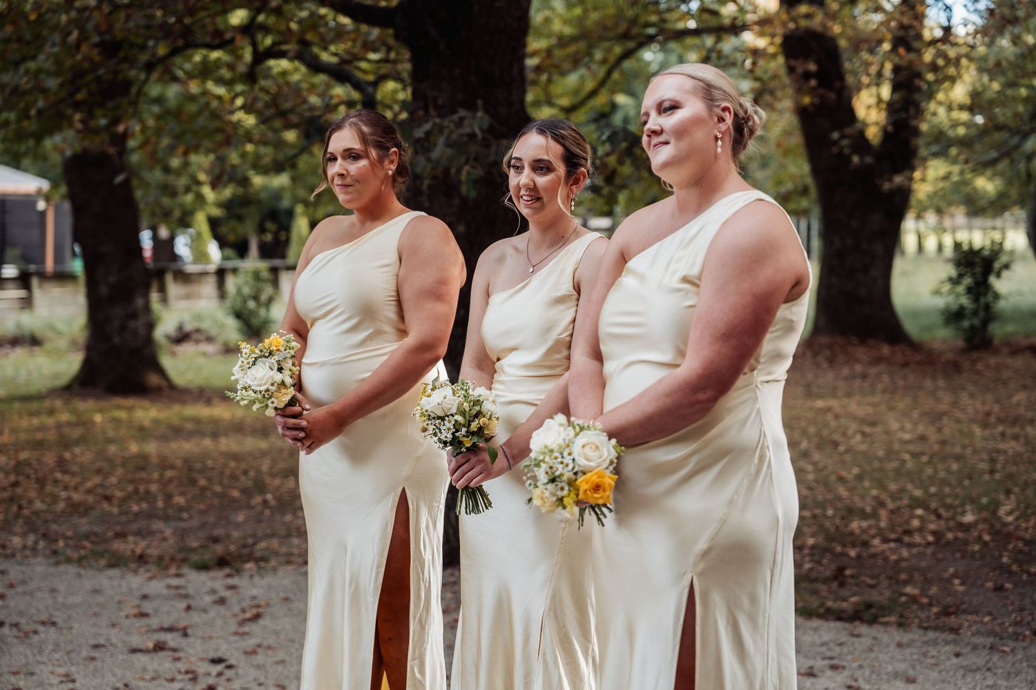 Three bridesmaids in ivory satin one-shoulder gowns holding yellow and white bouquets outdoors.