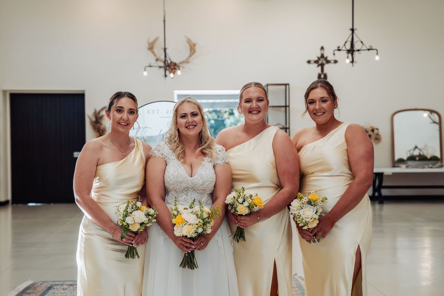 Bride poses with three bridesmaids in matching cream one-shoulder gowns holding yellow and white bouquets.