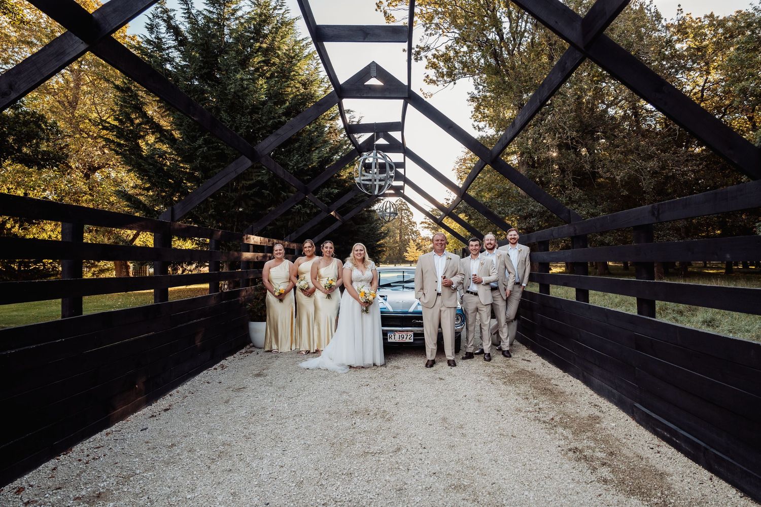 Wedding party poses together on a wooden bridge with a vintage truck in the background on a sunny day.