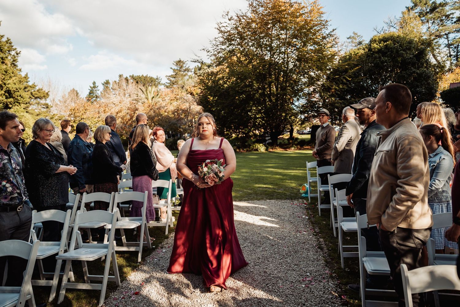 Bridesmaid in burgundy gown walks down outdoor wedding aisle lined with white chairs and guests.