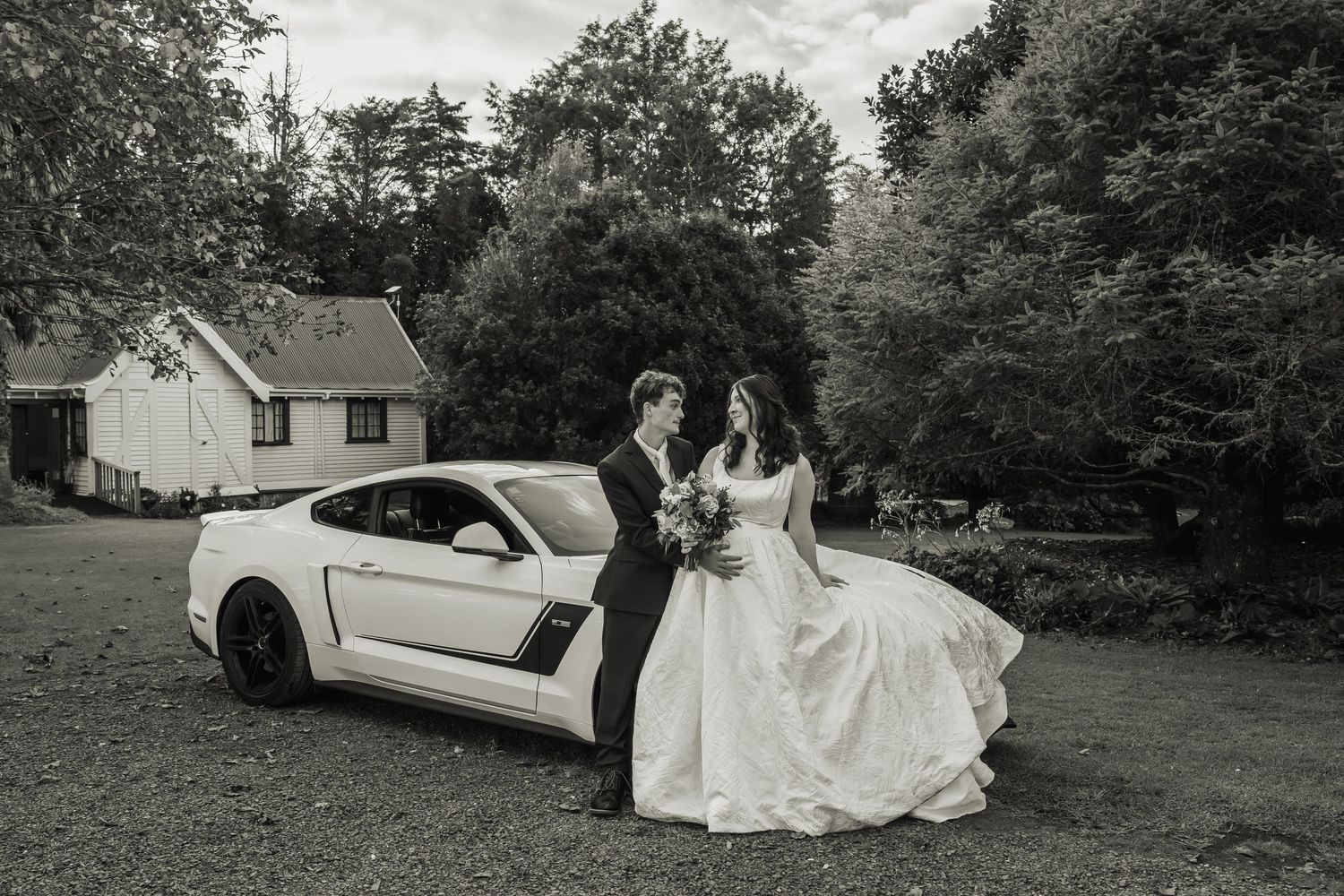 Black and white photo of bride and groom sharing romantic moment beside white Mustang.