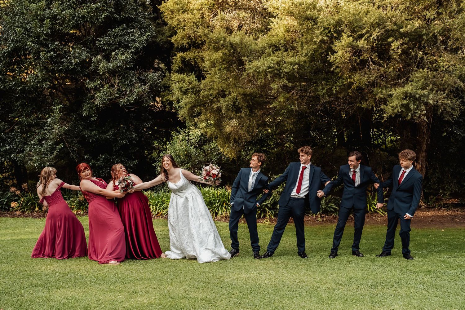 Fun wedding party photo with bridesmaids in burgundy dresses and groomsmen in navy suits on garden lawn.