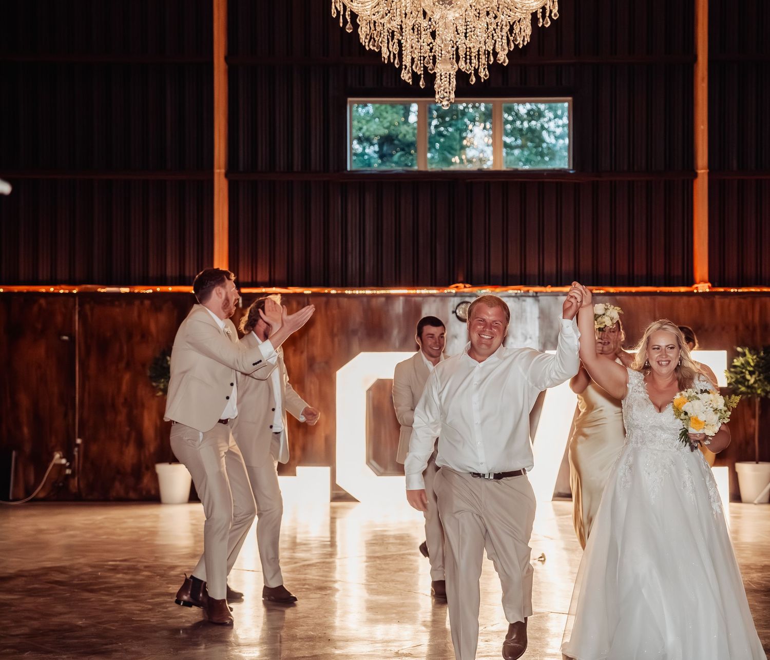 Bride and groom make grand entrance into rustic barn reception hall under crystal chandelier with cheering guests.