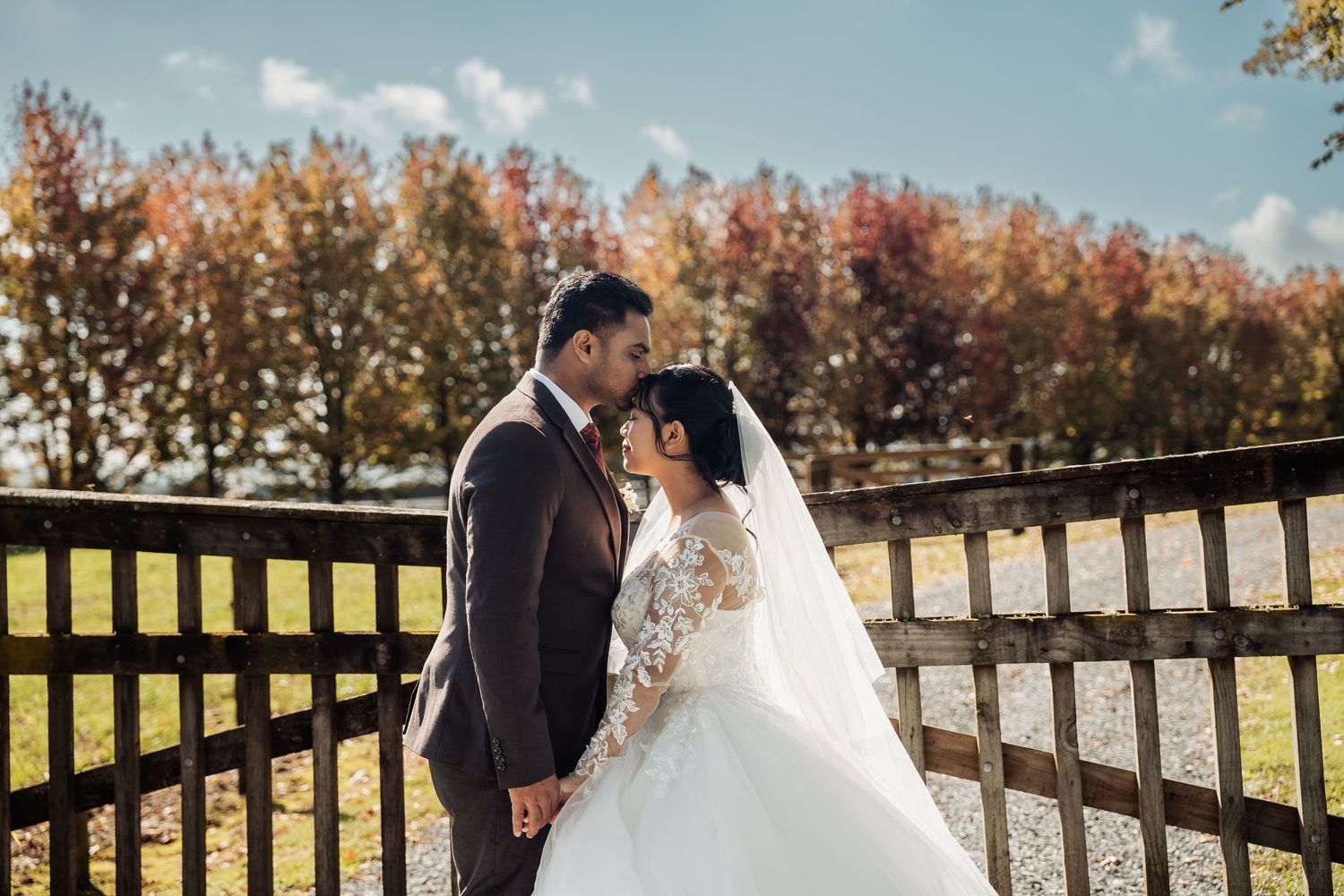Bride and groom sharing a tender kiss near a wooden fence with vibrant autumn foliage behind them.