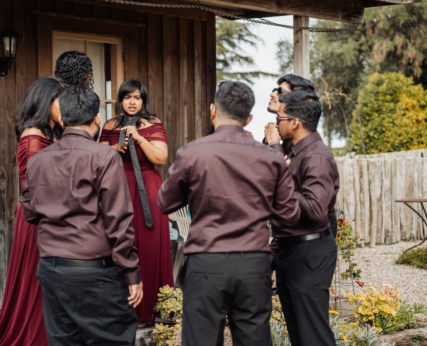 Wedding party in maroon attire gathering outdoors, groomsmen adjusting ties near a rustic wooden building.